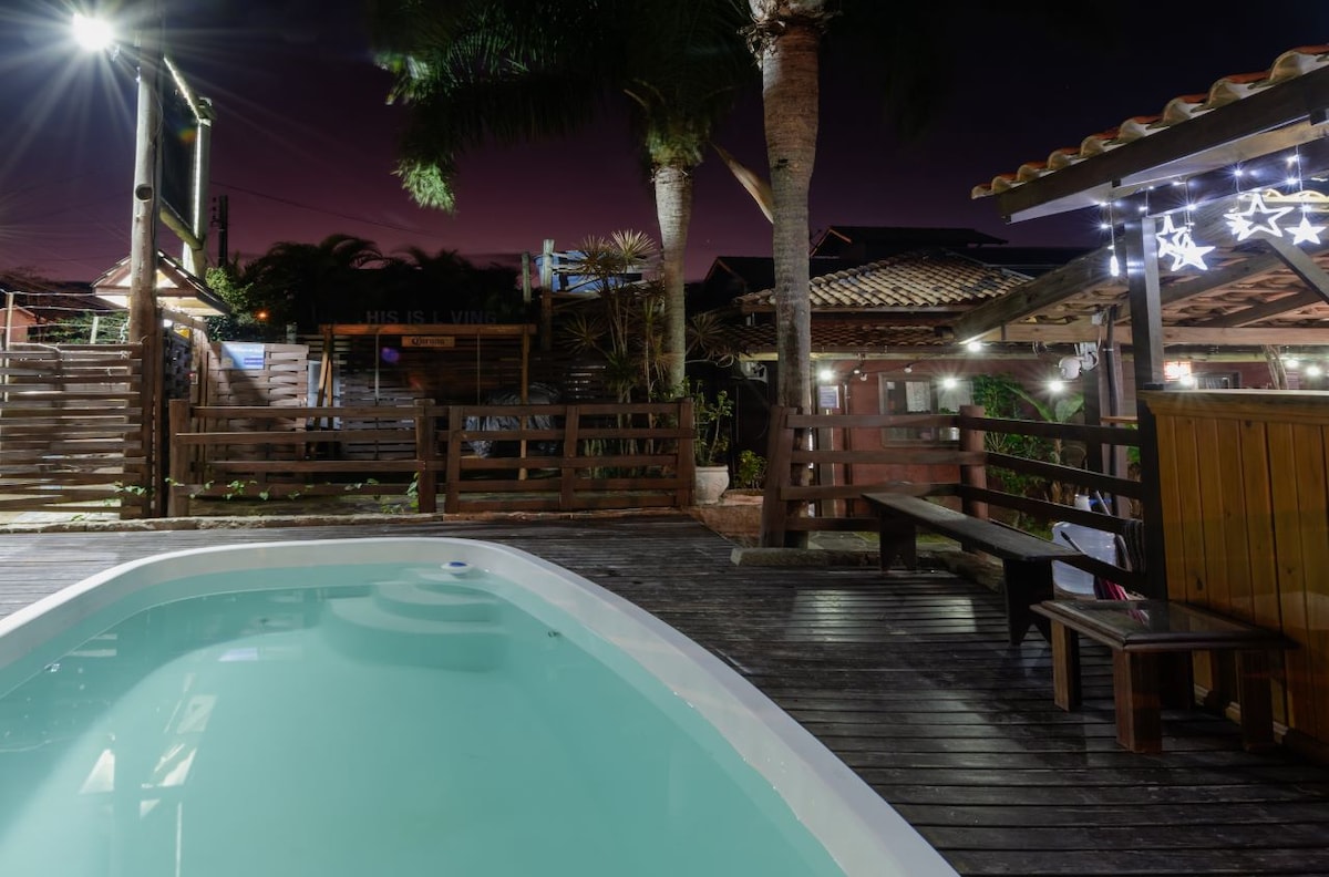 A tranquil night view of a pool surrounded by wooden decking and palm trees. Soft lighting enhances the inviting space, where seating arrangements are visible. Also seen are surrounding structures that provide a cozy ambiance.
