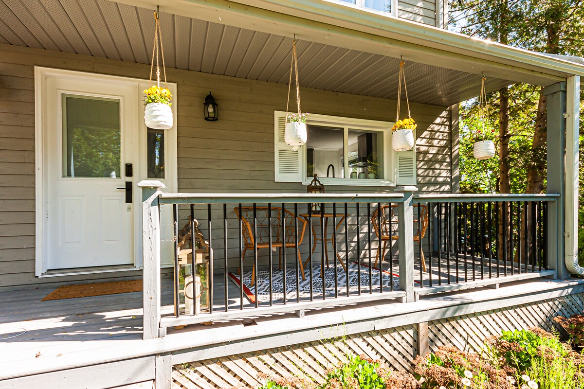 A welcoming porch features a grey wooden railing adorned with hanging planters filled with flowers. Comfortable wooden seating is arranged around a small table, inviting relaxation. Natural light is present, and a door leads into the cottage, enhancing the connection between indoor and outdoor spaces.