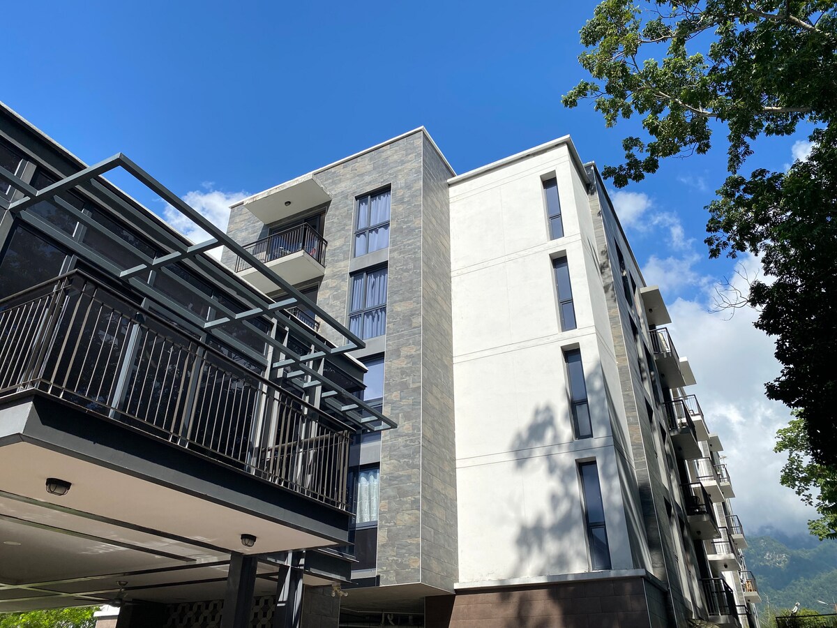 The exterior of a modern condominium building is captured under a clear blue sky. The structure features a combination of dark stone and light-colored walls, with large windows and a balcony on the second floor, offering a glimpse of the surrounding greenery.