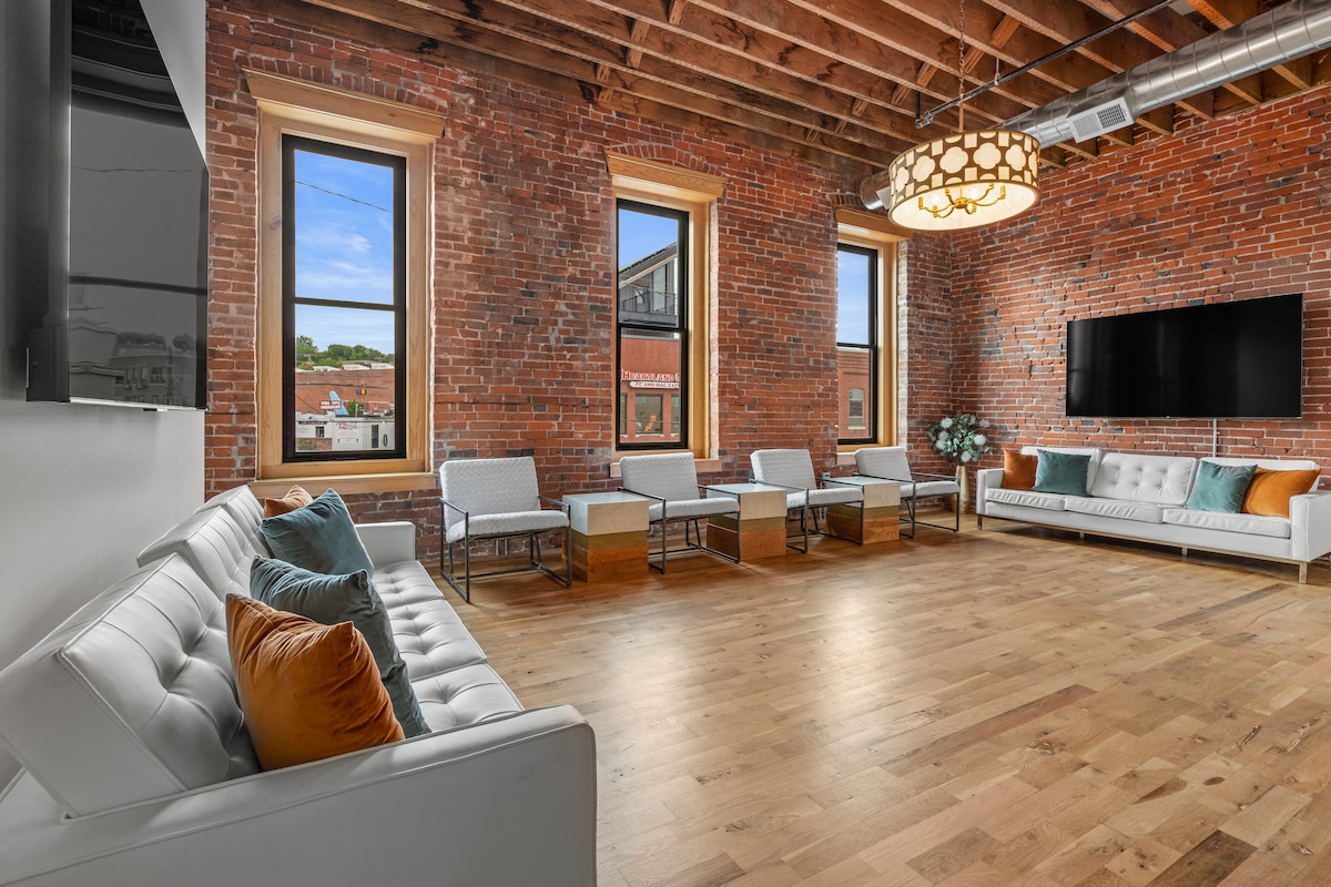 An open living area features high ceilings with exposed wooden beams, accentuated by large windows that allow natural light. Modern white furniture is arranged around a wooden coffee table, with decorative cushions adding color. A mounted flat-screen TV is visible on the exposed brick wall.