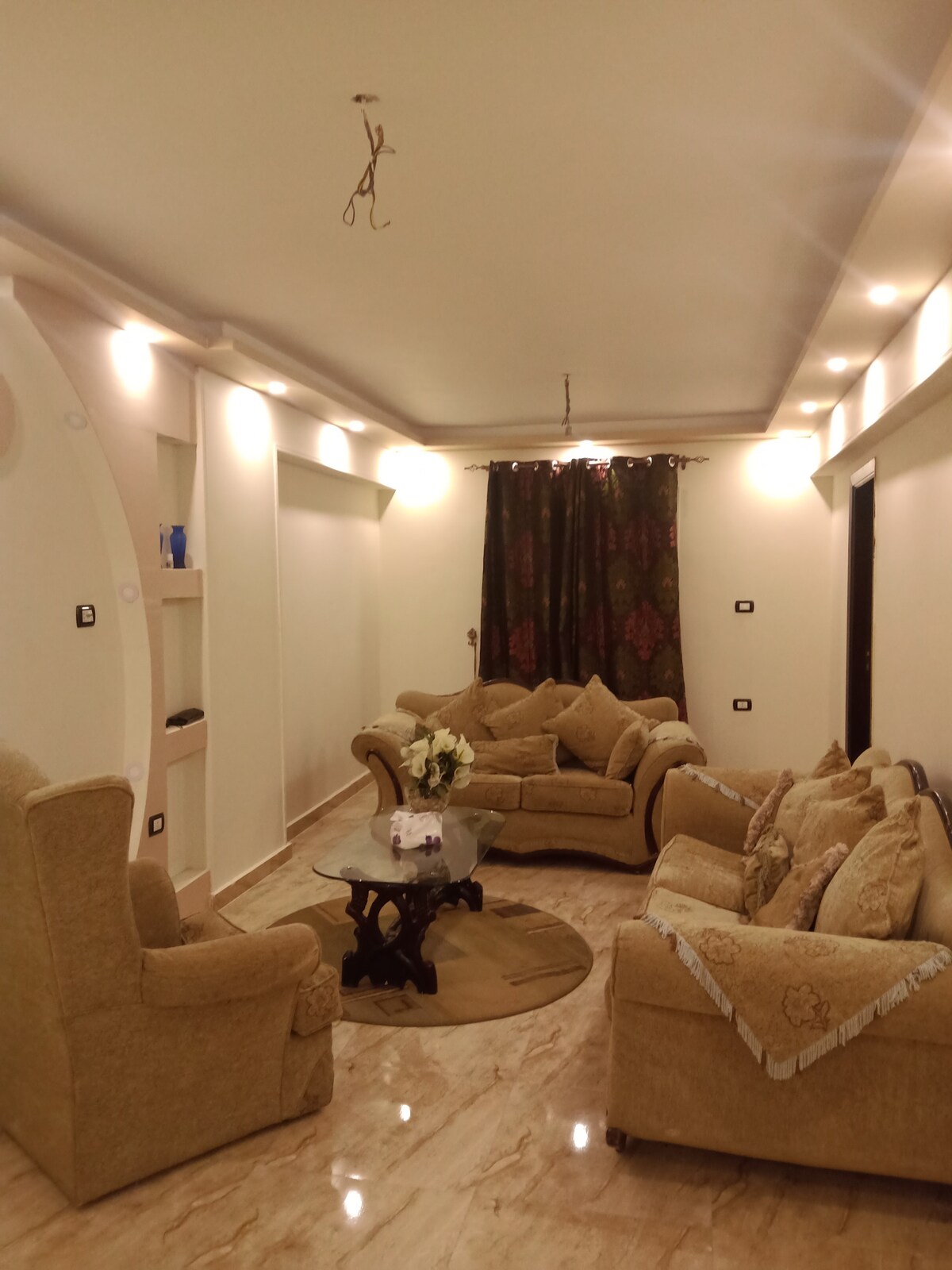 The living area features a collection of beige upholstered sofas arranged around a decorative glass coffee table. Soft lighting is provided by recessed fixtures and a chandelier, while dark curtains frame a window. A vase with flowers adds a touch of decor.