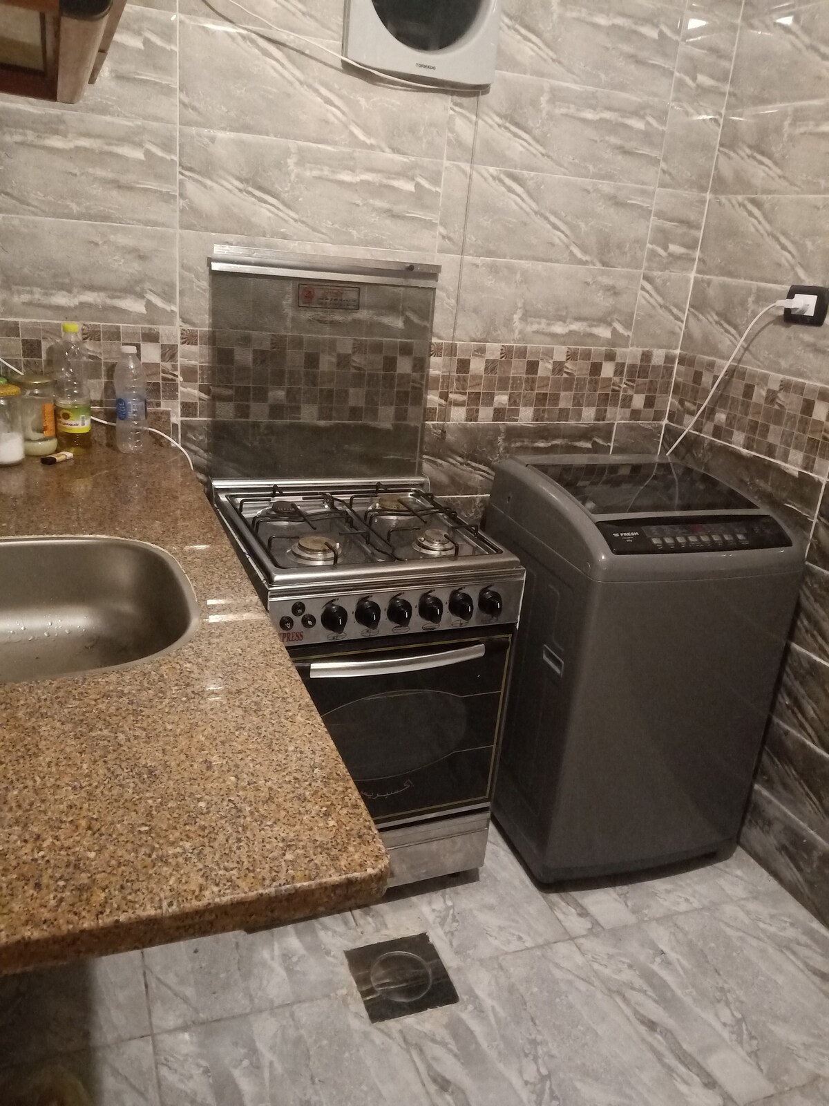 A compact kitchen area features a stainless steel gas stove with four burners and an oven below. A washing machine is positioned adjacent to the stove, while a granite countertop spans the length of the space. Mosaic tile accents add a decorative touch to the wall.