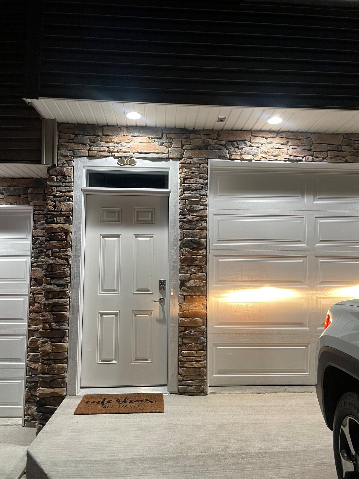 The entrance features a solid white door set against a textured stone facade. A welcoming doormat is placed on the concrete steps leading to the door, while motion-activated lights illuminate the area, enhancing visibility during evening hours.