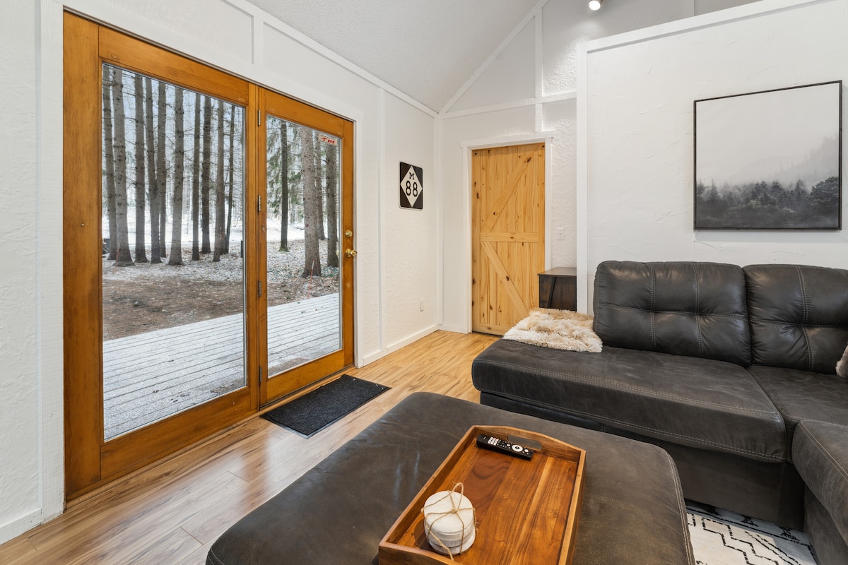 The living area features large glass doors offering a view of the serene forest beyond. A spacious sectional sofa is positioned opposite a wooden coffee table, enhancing the cozy setting. Natural light brightens the room, creating a welcoming atmosphere.