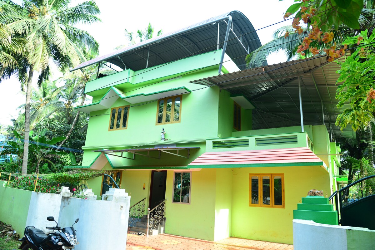 A two-story green house is surrounded by lush foliage. The building features large windows that allow natural light to fill the space. A covered porch area is present at the entrance, complemented by a tiled walkway. A motorcycle is parked nearby.