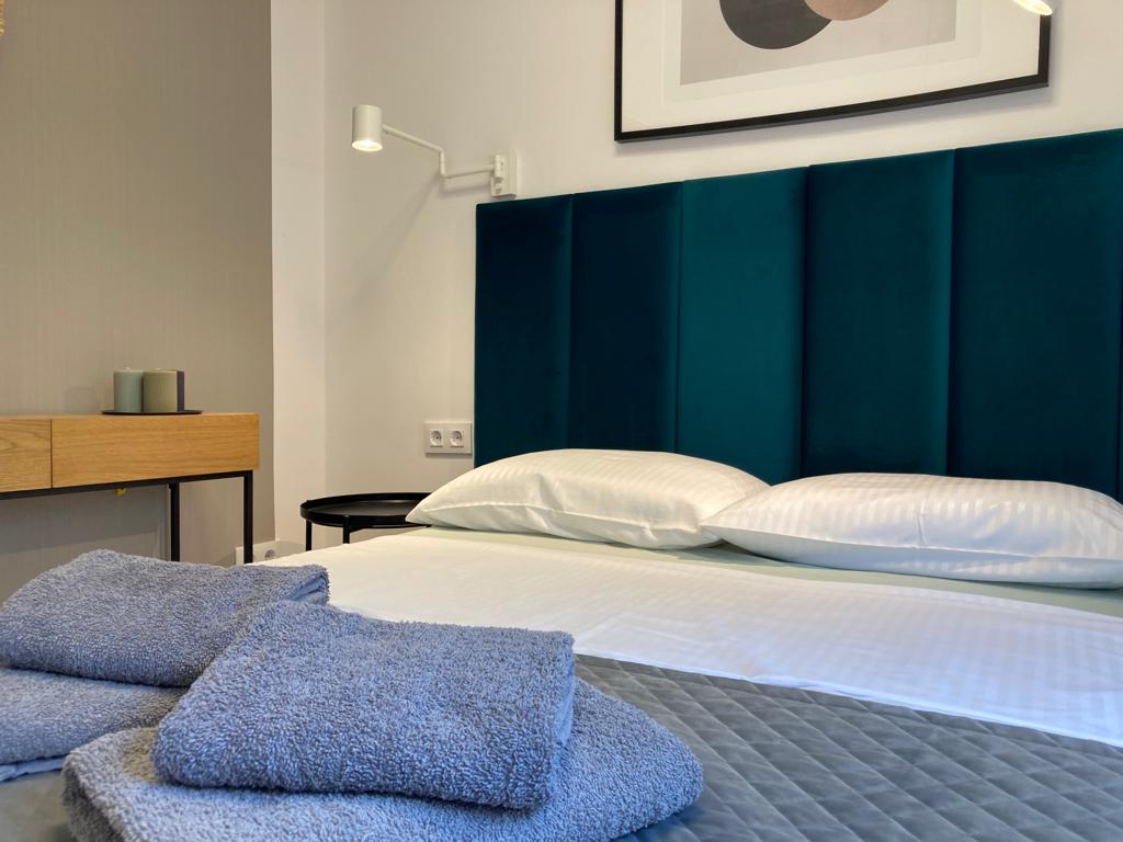 A minimalistic bedroom features a spacious bed with a dark green headboard and white bedding. Soft, folded towels are placed on the bed. A small wooden dresser and a side table complement the room, with subtle wall art adding a touch of elegance.