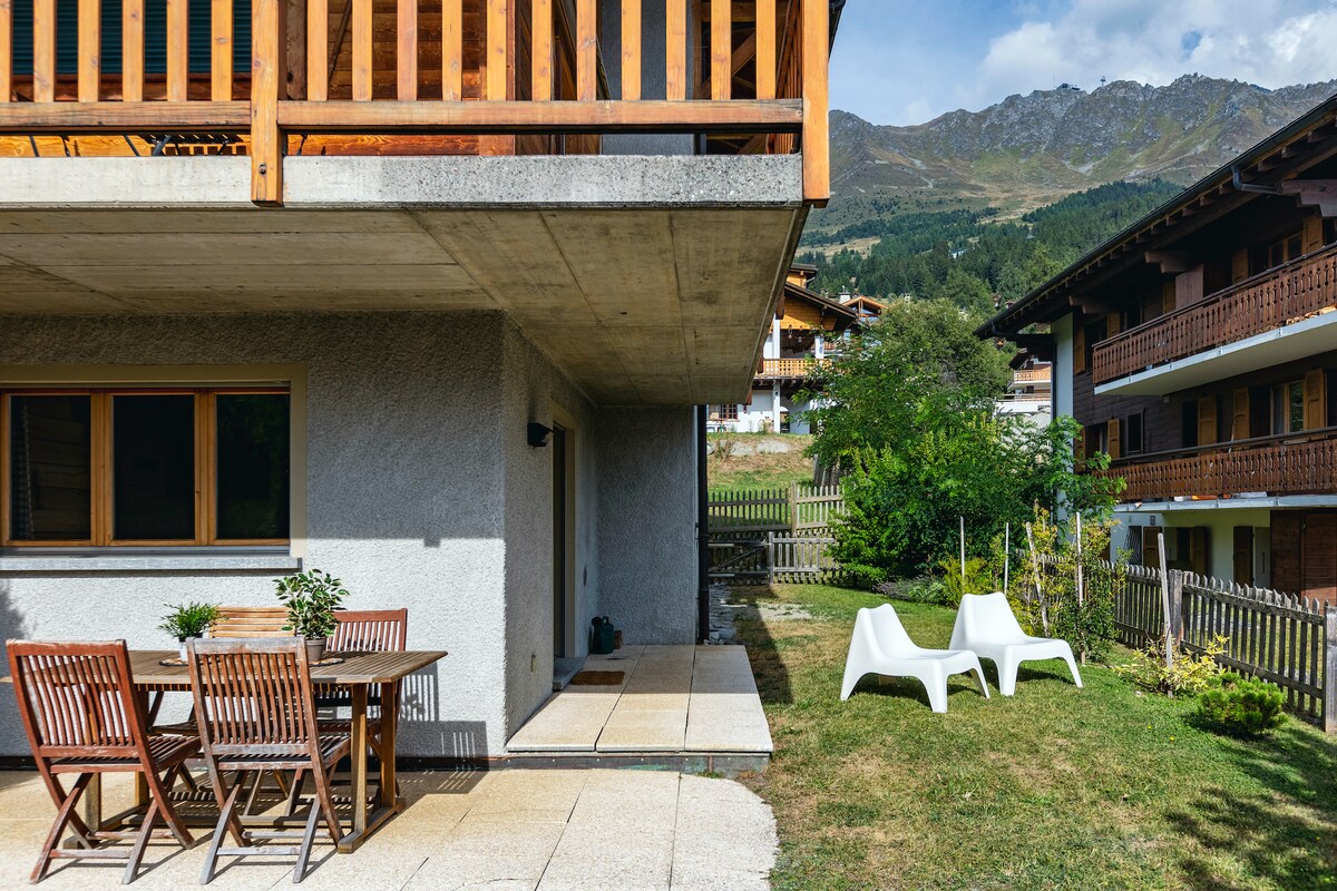 An outdoor terrace features a wooden dining table surrounded by slatted chairs. Nearby, two white lounge chairs are situated on a grassy area. A low stone wall and wooden fencing create a boundary, with mountainous scenery visible in the background.