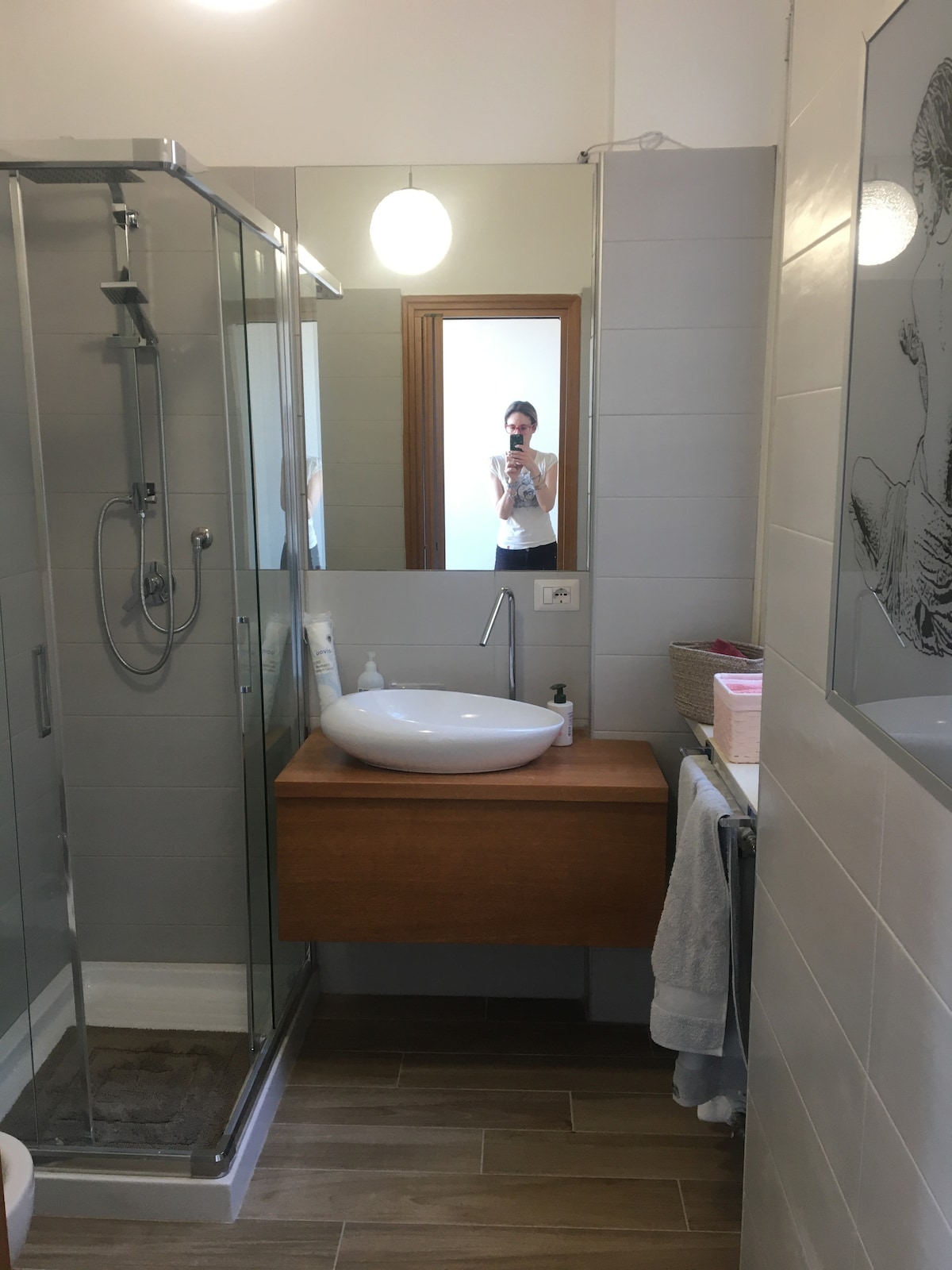 The bathroom features a spacious walk-in shower enclosed with glass, a modern circular sink on a wooden vanity, and a large mirror reflecting the room. Soft lighting is provided by a hanging fixture, and neutral-tone tiles enhance the clean design.