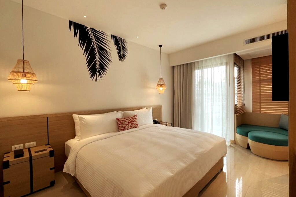 A modern studio features a spacious bed adorned with crisp white linens and a decorative pillow. Light fixtures with woven details hang above the bedside tables. Large glass doors lead to a quiet balcony area, complemented by a cozy seating nook bathed in natural light.