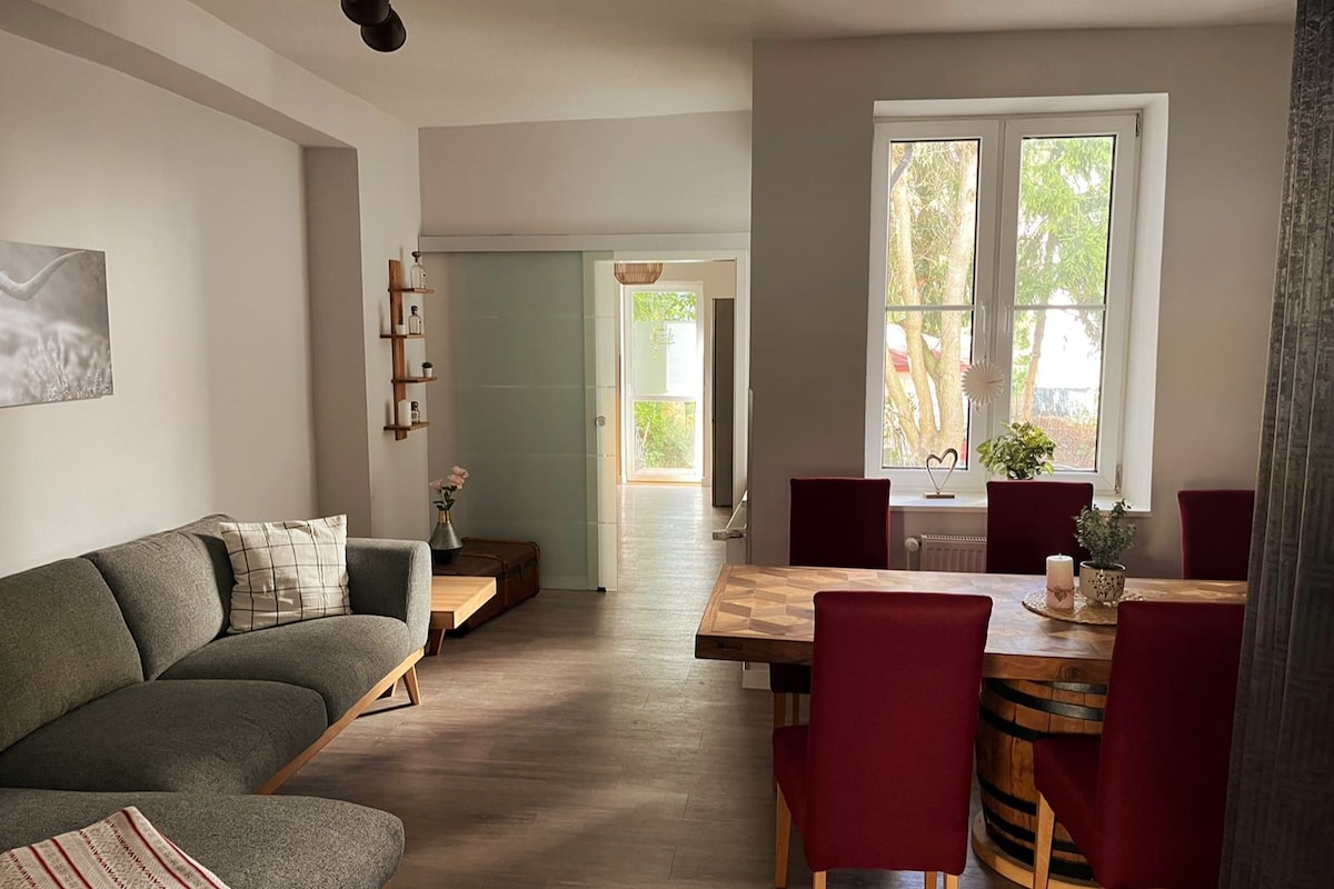 A bright living area features a comfortable sectional sofa, inviting relaxation. A wooden dining table with red chairs is positioned nearby. Natural light streams through large windows, illuminating the space, while an entryway leads to a glimpse of the outdoor area.