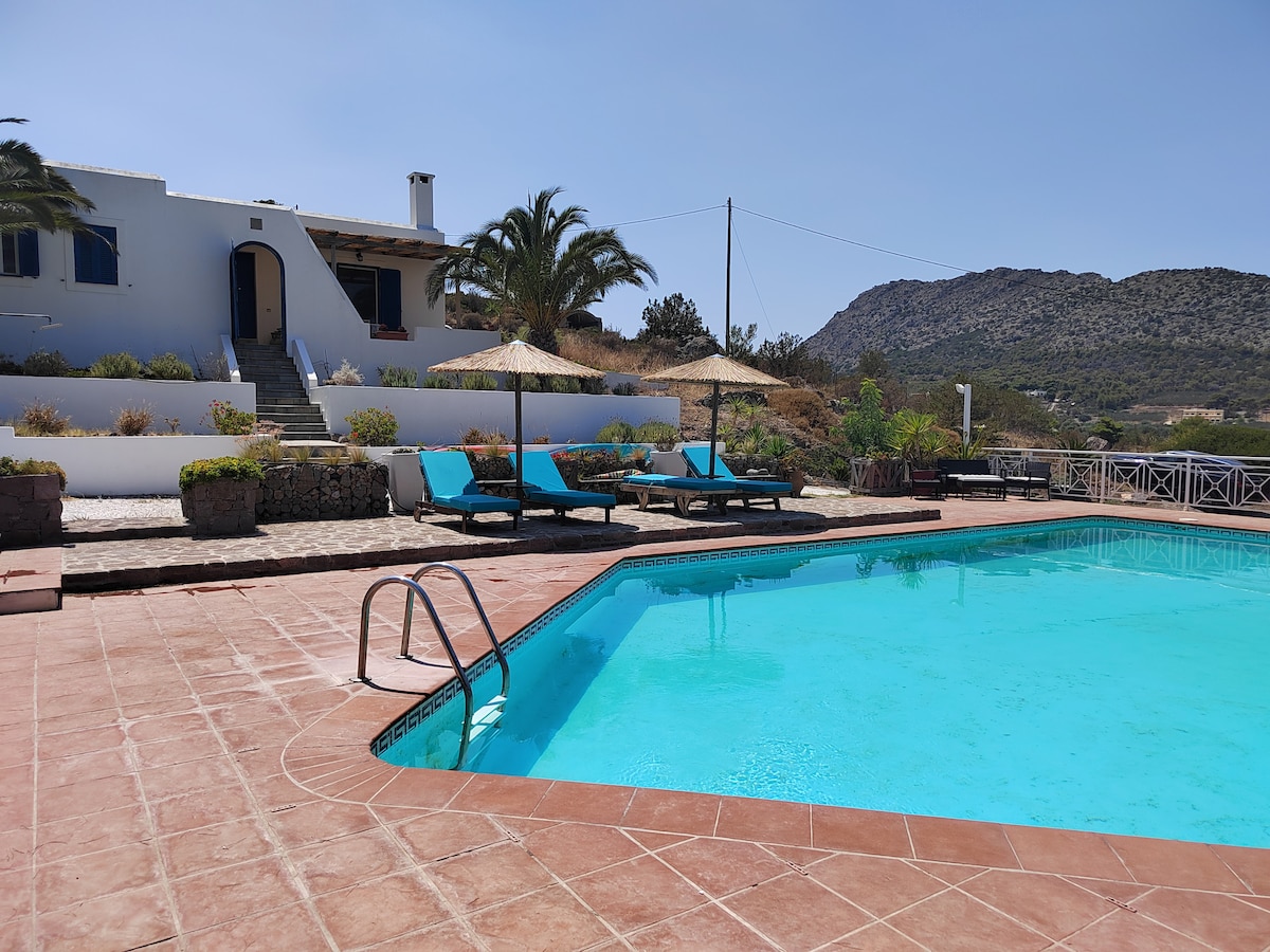 A refreshing swimming pool is surrounded by sun loungers and parasols, offering relaxation in the sun. The property is visible in the background, bordered by lush greenery and distant hills, contributing to a serene atmosphere.
