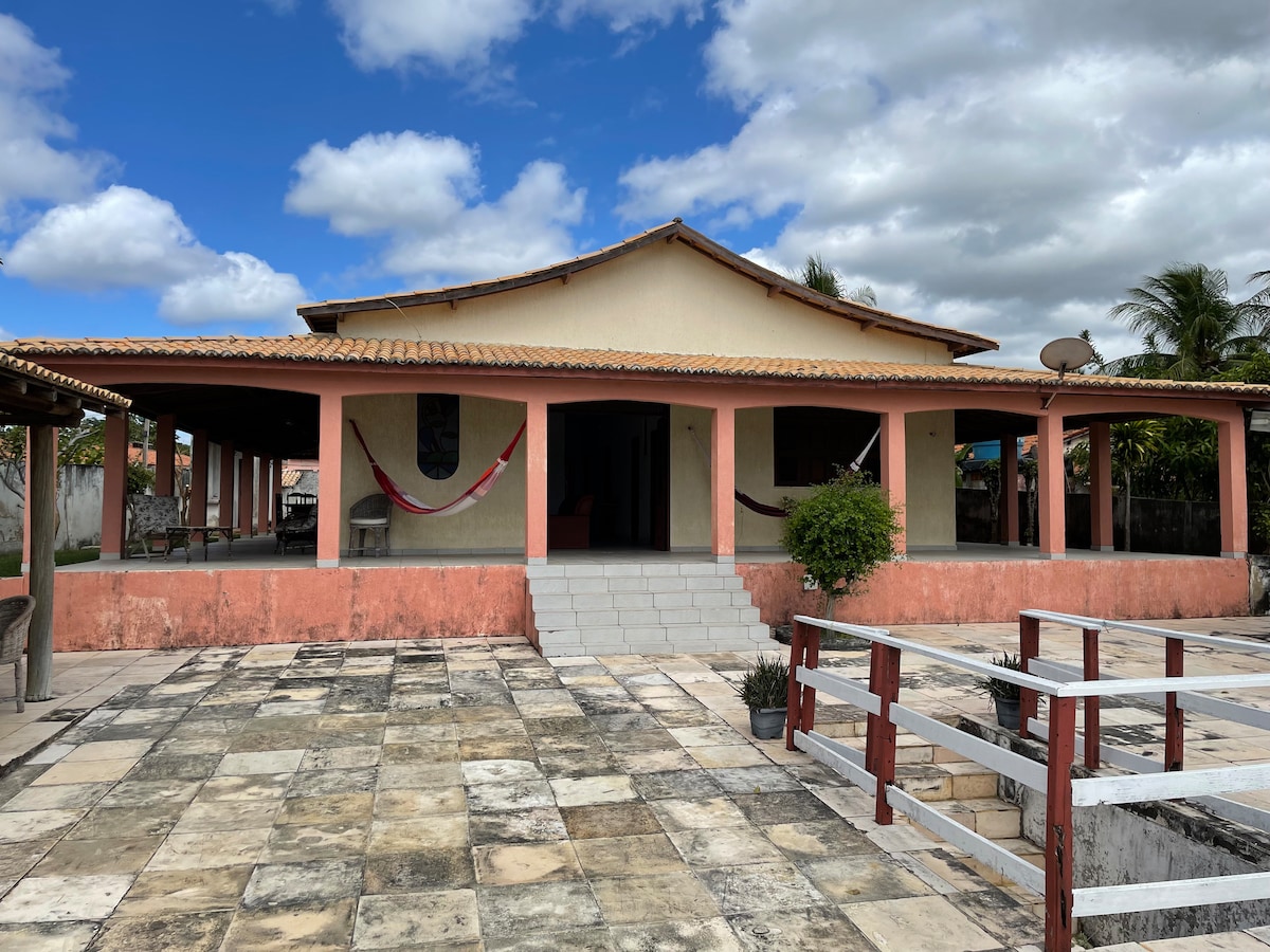 A spacious house features a large, covered porch with several hammocks hanging from the pillars. The exterior showcases terracotta accents against a pale yellow facade, with a well-maintained stone patio and a walkway leading to the entrance. Lush greenery surrounds the property, complemented by a clear blue sky.