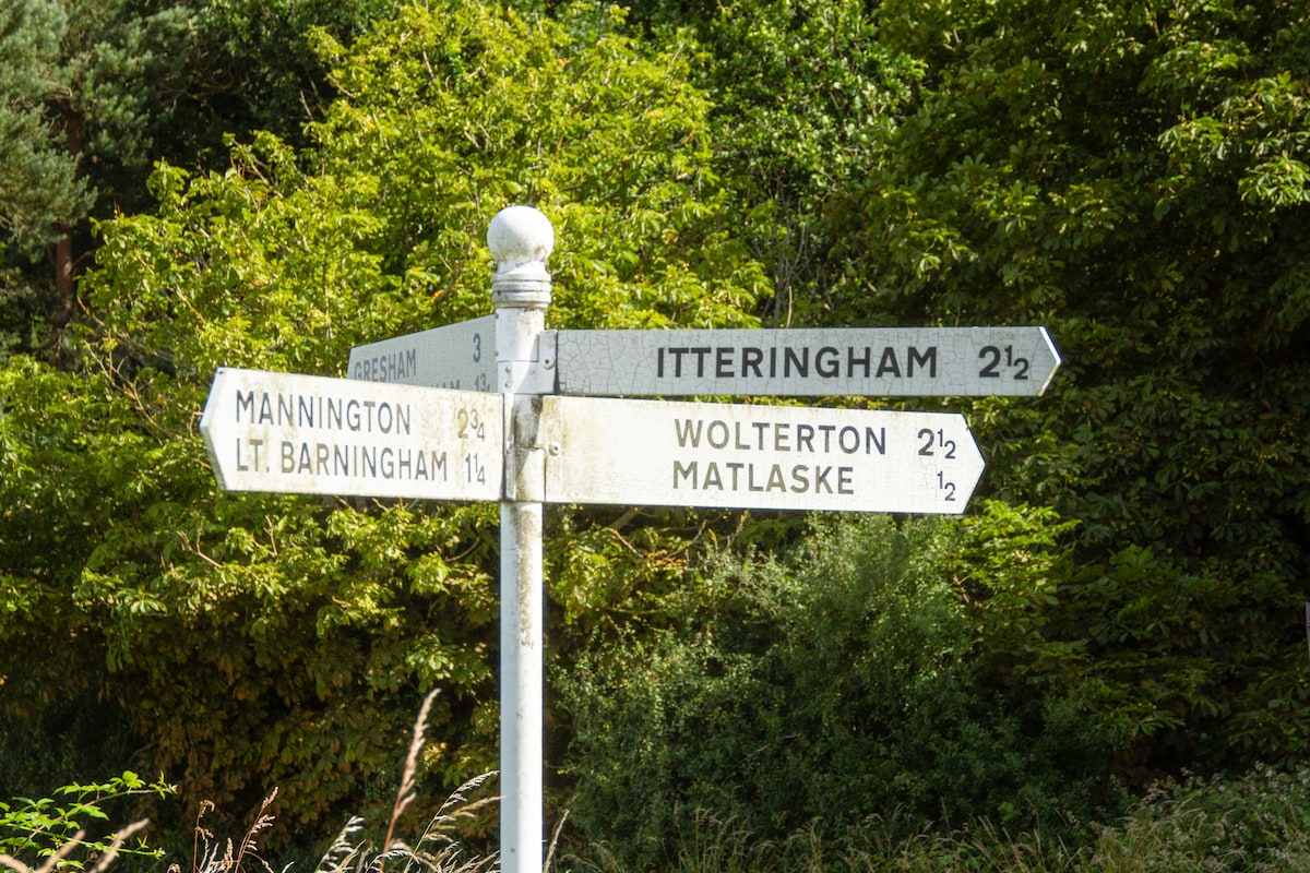 A weathered directional signpost is displayed, pointing toward various destinations including Wolterton and Mannington. The sign's arms show distances in miles, with a rustic design complementing the lush greenery in the background, enhancing the rural setting.