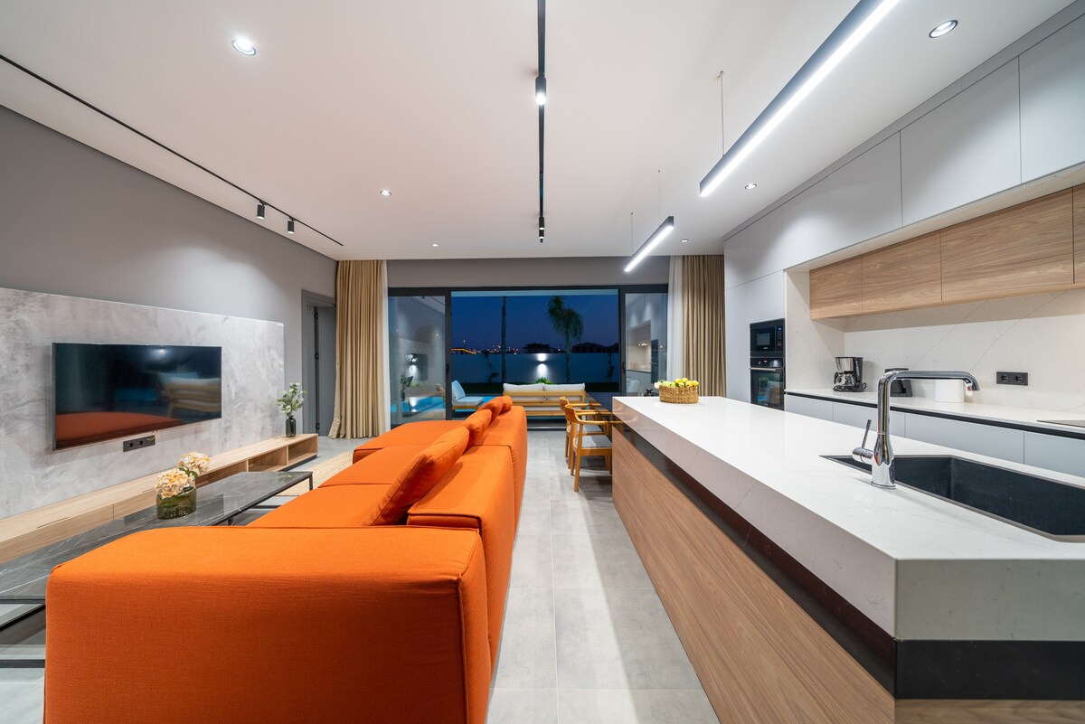 A modern living area is showcased, featuring an orange sofa that faces a wall-mounted television. A kitchen island with a sleek countertop is visible, alongside wooden dining chairs set around a table. Large glass doors provide a view of the outdoor space.