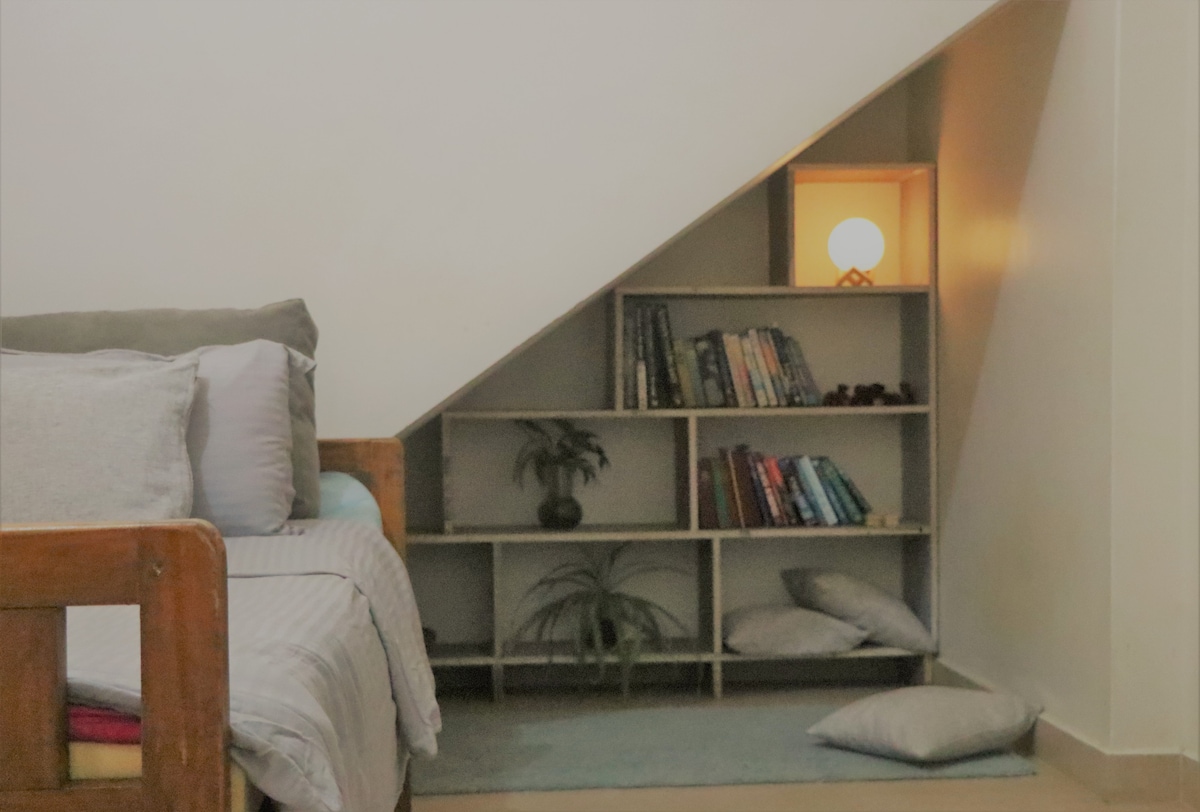 A cozy reading nook is presented beside a spacious bed, featuring a mini library on a wooden shelf against the wall. Soft cushions are arranged on the floor, and a warm light illuminates a decorative plant and a collection of books.