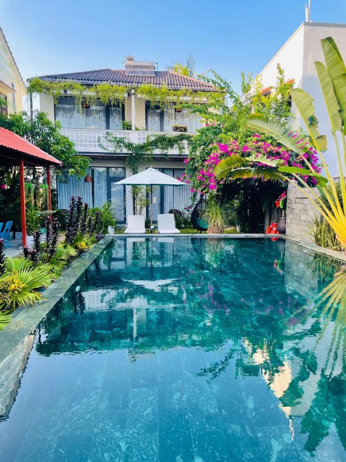 A serene outdoor swimming pool is surrounded by lush greenery and vibrant flowers. Comfortable lounge chairs are positioned under a white sunshade. A multi-story building with a verdant balcony and decorative plants is seen in the background against a clear blue sky.