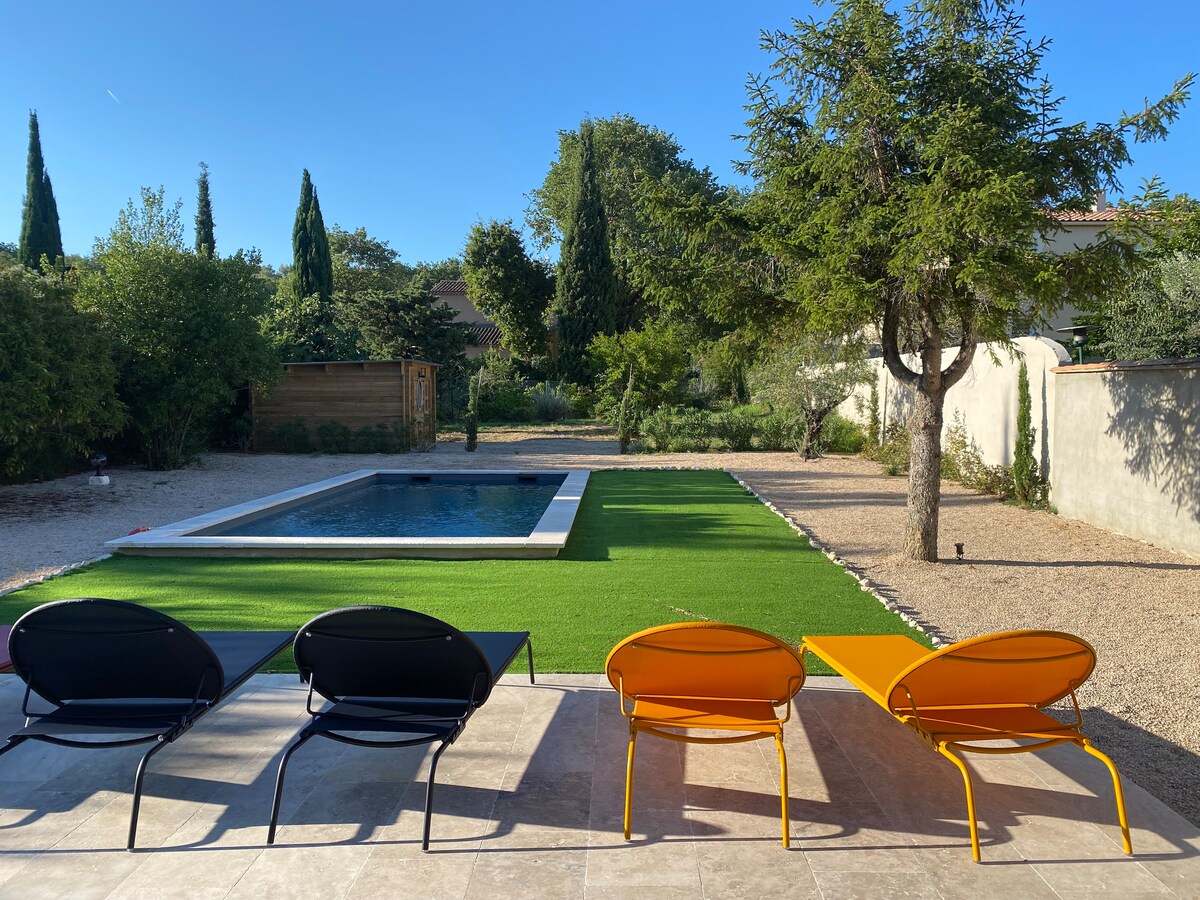 Four lounge chairs in black, orange, and yellow are positioned on a stone patio, overlooking a landscaped garden. A rectangular pool is visible, surrounded by manicured grass and natural greenery, with tall trees providing shade in the background.
