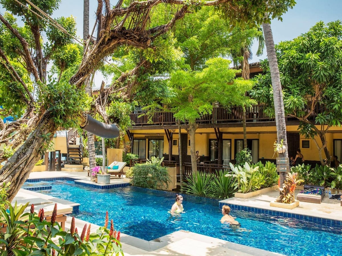 A serene swimming pool is surrounded by lush greenery and tropical trees. Guests are seen enjoying the water, while sun loungers and vibrant plants enhance the tranquil setting. The resort's buildings provide a colorful backdrop with multiple levels and spacious balconies.