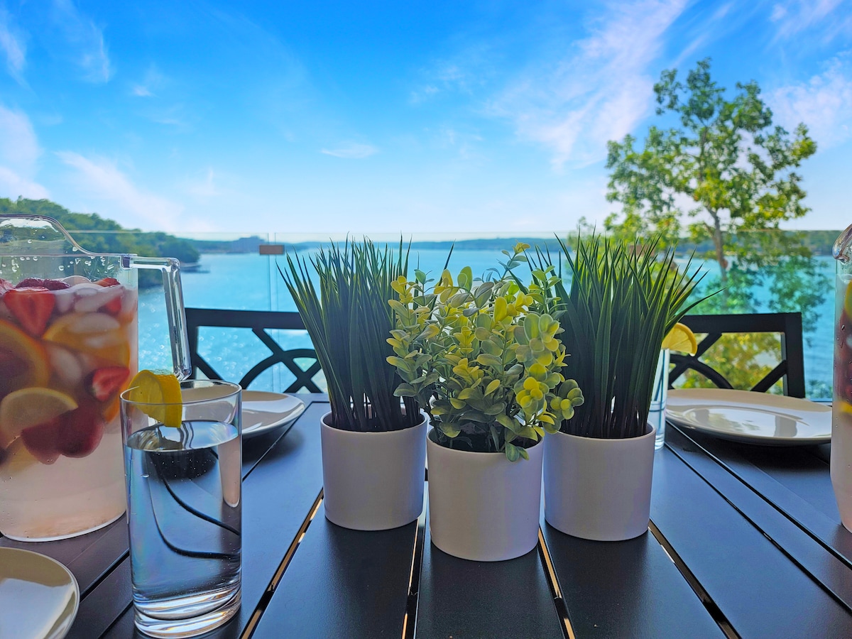 A dining table is set against a backdrop of serene water with a clear blue sky. Centered are three small potted plants and glasses of water, while a pitcher with fruit-infused drink is visible to the side, complementing the inviting outdoor dining experience.