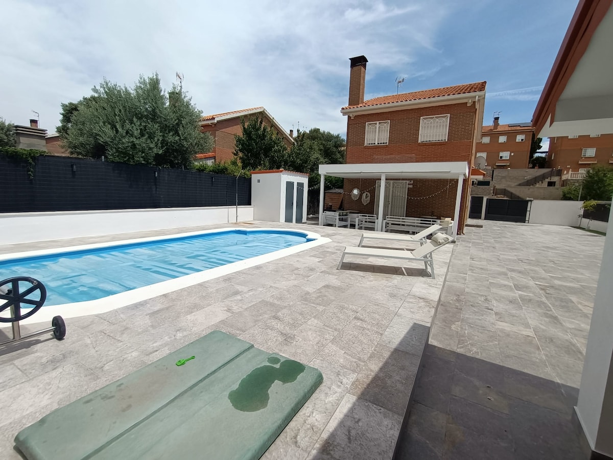 A rectangular swimming pool is surrounded by a spacious patio featuring loungers and a dining area. Lush greenery borders the property, and a modern structure is visible in the background. The area offers a blend of leisure and comfort, with sunlight illuminating the outdoor space.
