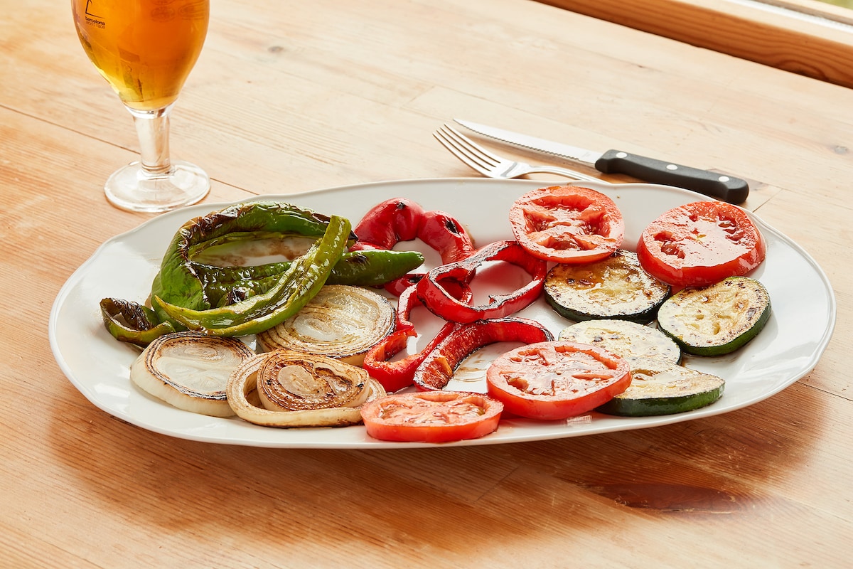 A large, oval platter displays a vibrant assortment of grilled vegetables, including green and red peppers, zucchini, onions, and tomatoes. The vegetables are arranged artfully, showcasing their colors and textures. A glass of beer and a knife are visible in the background, resting on a wooden table.
