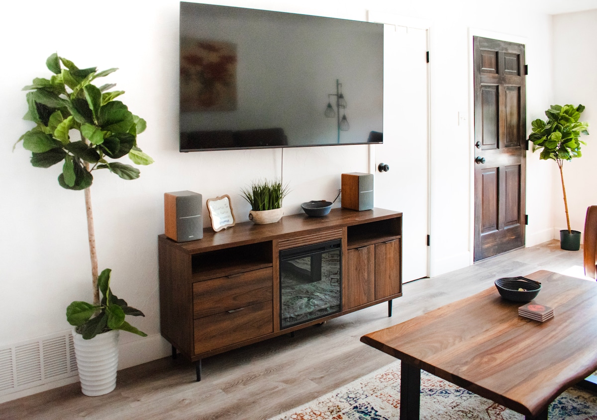 A modern entertainment center with a large flat-screen TV mounted above a wooden unit is displayed. The unit contains various compartments and speakers. Lush potted plants are positioned on either side, complementing the light and airy decor. A dark wood door is partially visible in the background.