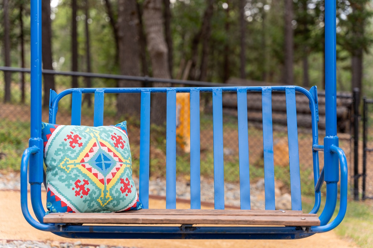 A blue swing set is displayed in a tranquil outdoor area, surrounded by trees. A brightly patterned cushion adds a pop of color to the wooden seat, providing a cozy touch to this inviting space.