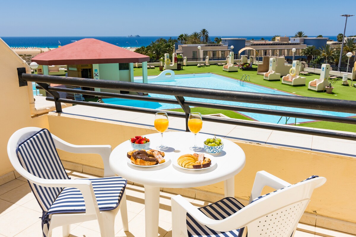 A terrace set for a meal features a white table with two chairs, holding a spread of pastries, fruits, and drinks. In the background, a swimming pool and palm trees are visible, with the ocean glistening under the sun.