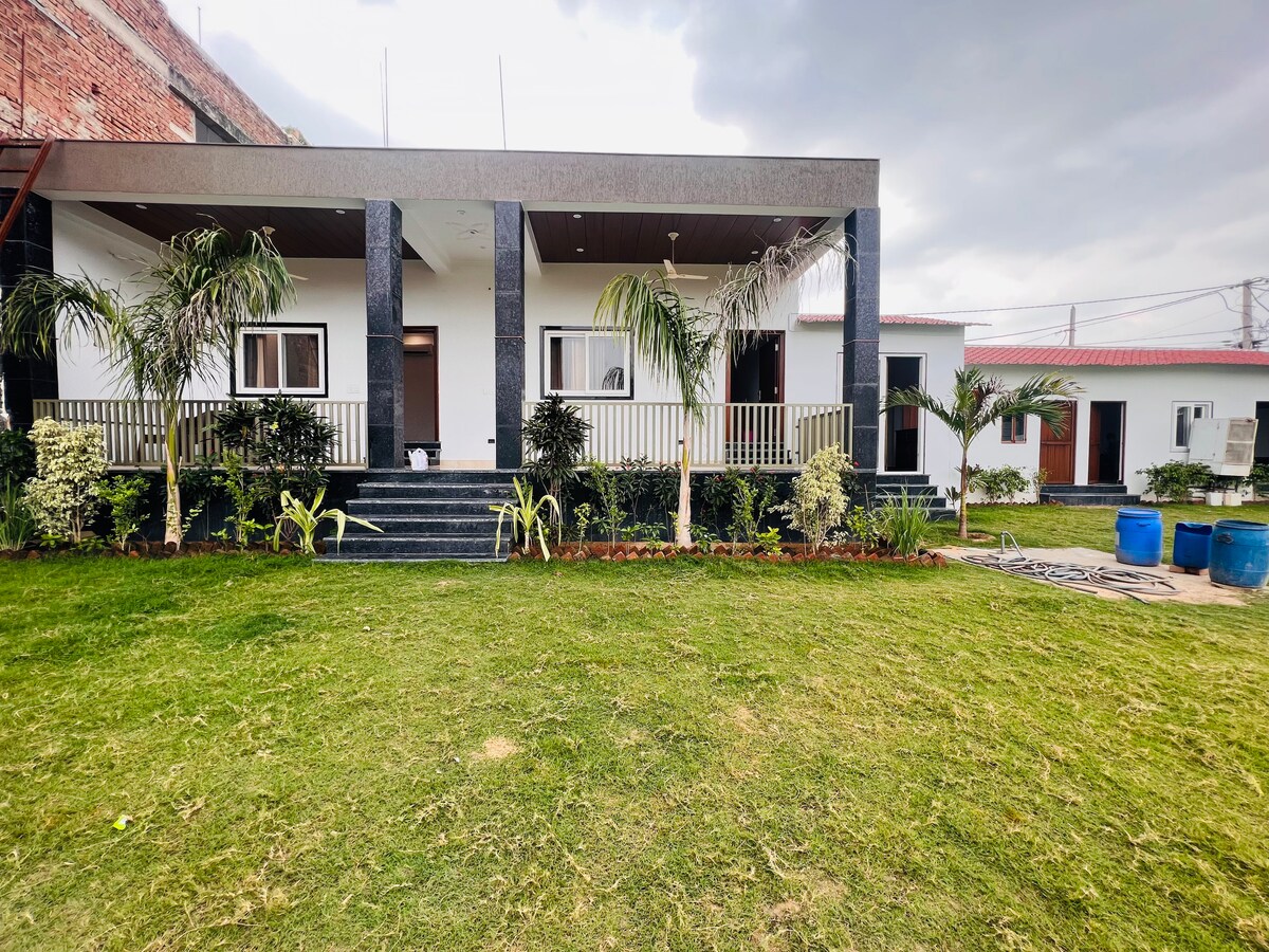 A modern exterior of a property is shown, surrounded by lush green grass and ornamental plants. The facade features a spacious porch with support columns and a staircase leading to the entrance. Blue containers are visible in the garden area, contributing to the serene outdoor environment.