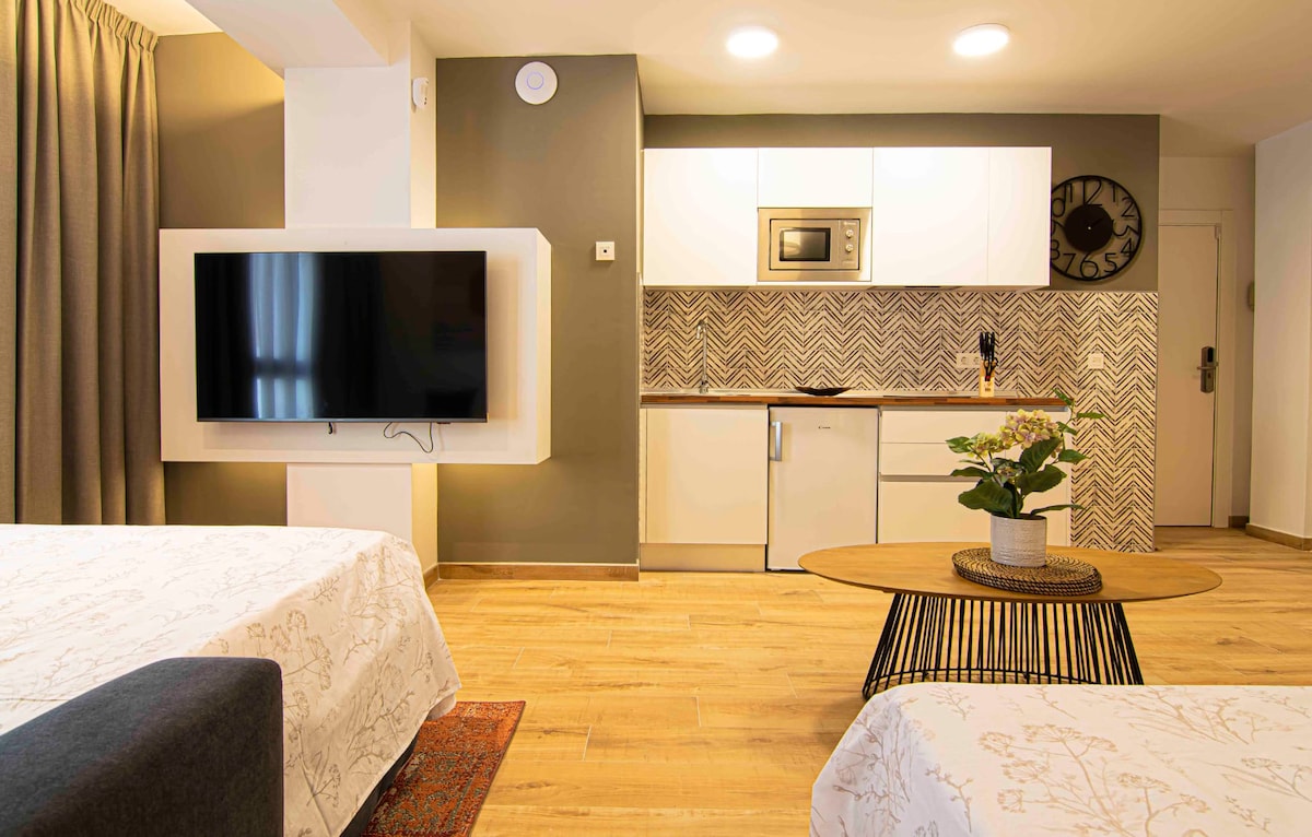 A modern living space is displayed, featuring two spacious beds positioned beside a circular coffee table. A flat-screen television is mounted on the wall, opposite a kitchenette equipped with a microwave and refrigerator. Light wooden flooring complements the soft tones of the decor.