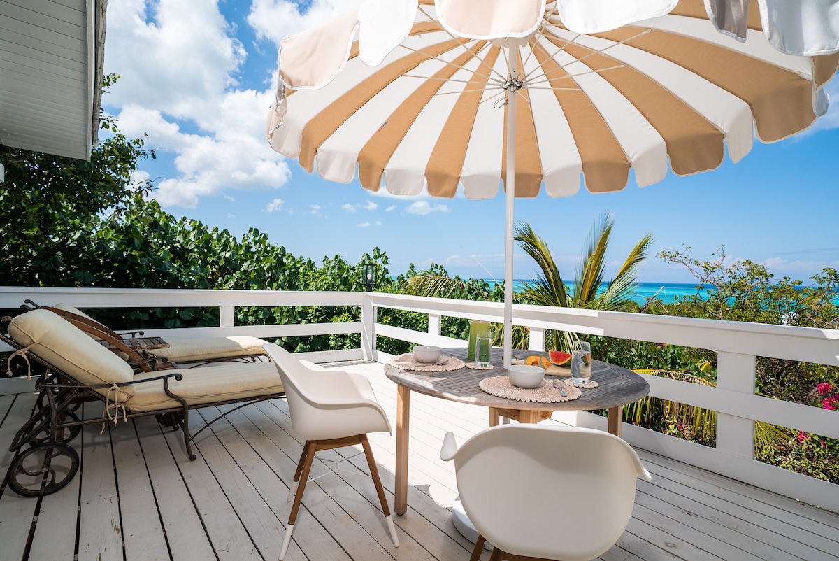 An outdoor dining area is featured on a private terrace, showcasing a round table with white chairs and a striped umbrella providing shade. A reclining lounge chair is positioned nearby, with lush greenery and distant ocean views enhancing the serene setting.
