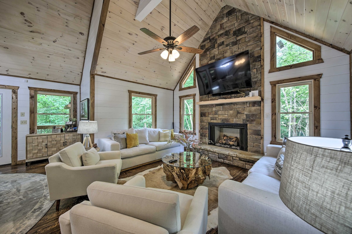 A spacious living area is highlighted by tall ceilings and a stone fireplace. Comfortable seating is arranged around a unique coffee table made from natural wood. Large windows allow for ample natural light and views of the surrounding greenery.