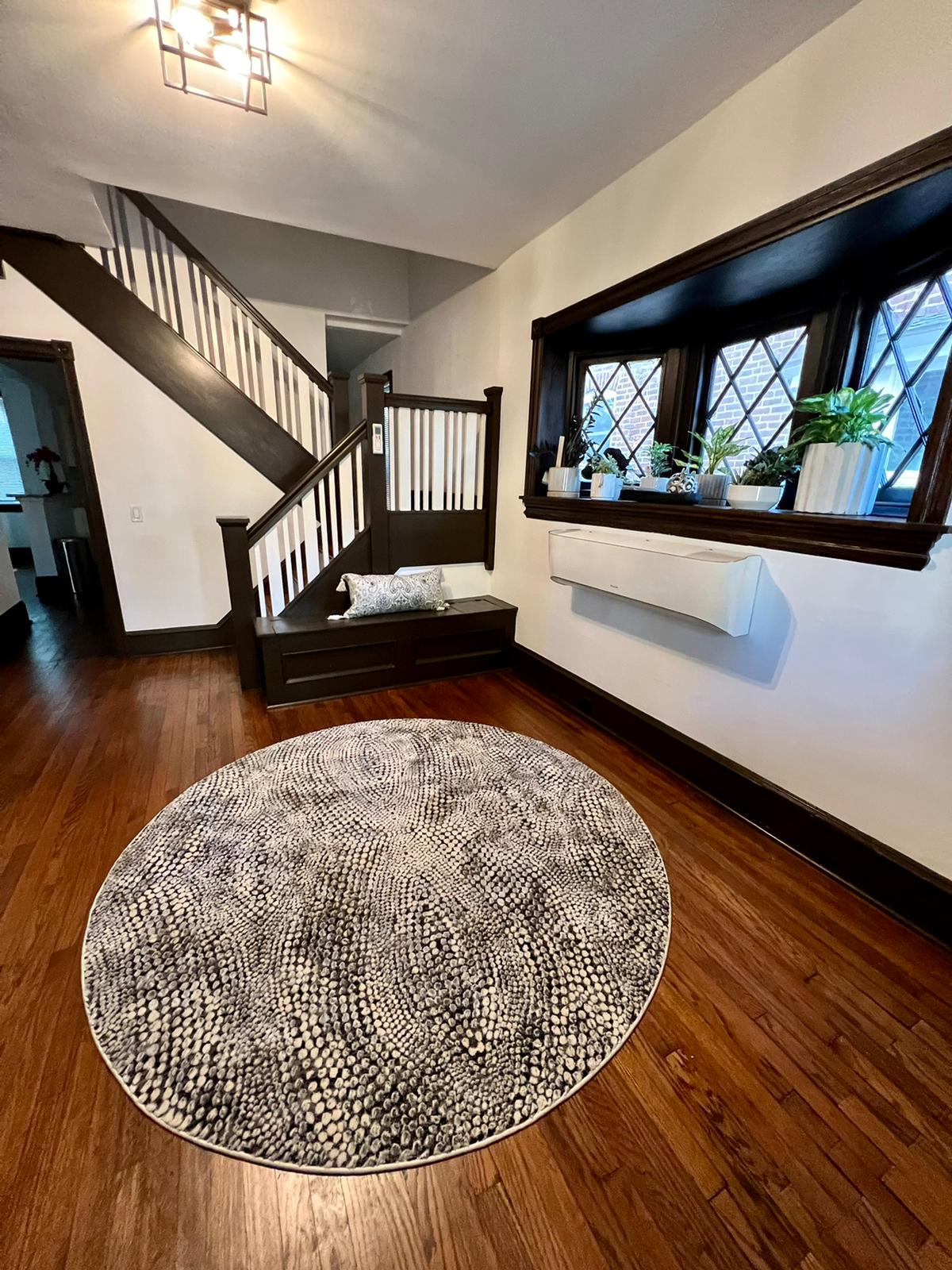 A welcoming entryway features polished hardwood floors and an oval area rug with a textured design. A comfortable bench provides seating under a window adorned with potted plants. The staircase, leading to the upper levels, offers a view of the intricate railing and patterned window panes.