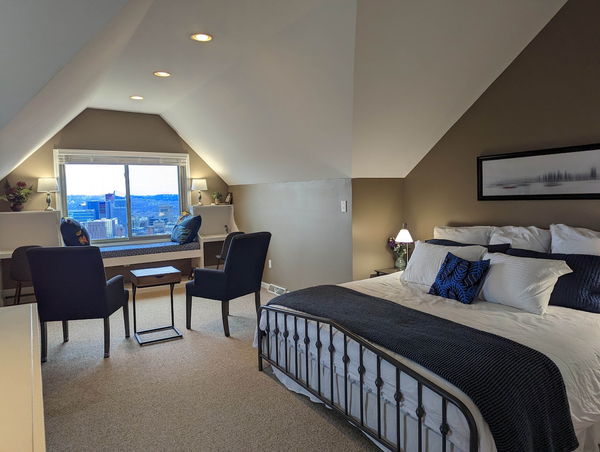 A spacious loft suite features a king bed with decorative pillows, beside tables with lamps, and seating area with two chairs and a table. Large windows provide an unobstructed view of downtown Pittsburgh, allowing natural light to enhance the neutral tones of the room.