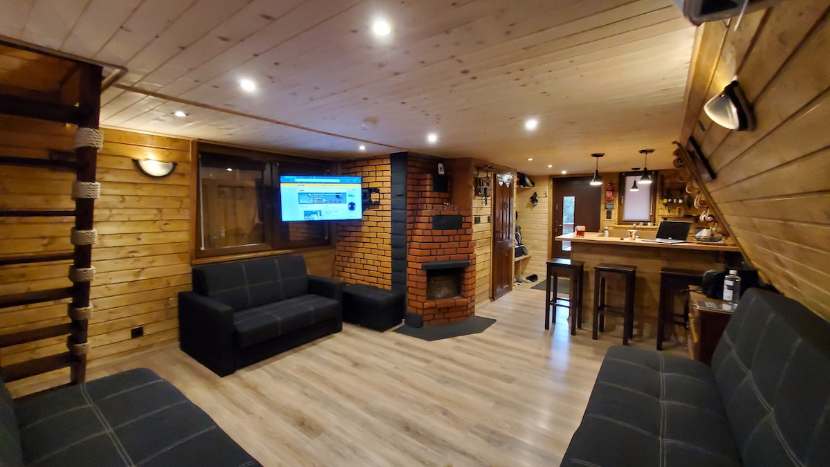 The interior space features wooden walls and a cozy seating area with two dark sofas positioned around a central fireplace. A television is mounted on the wall, and a bar area is visible in the background, complemented by high stools and warm lighting.