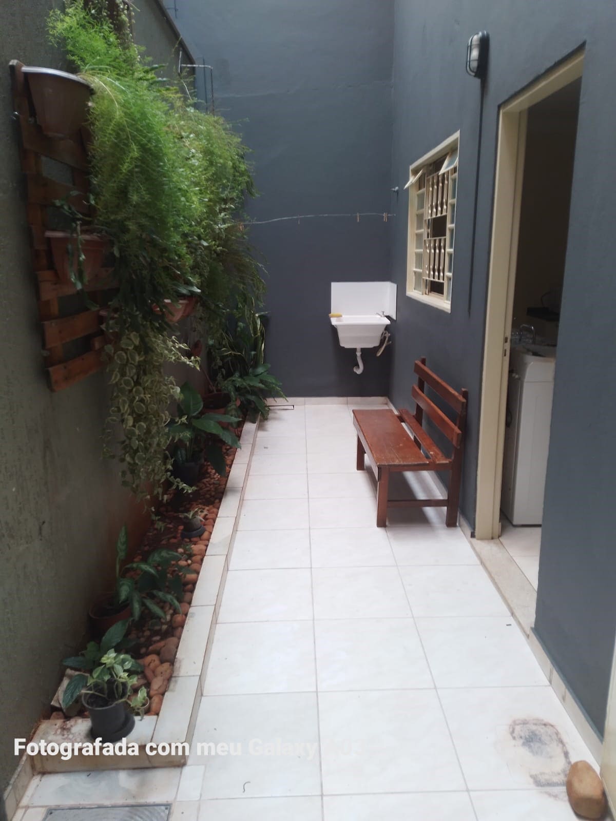 A narrow corridor is lined with green plants along the walls, creating a sense of freshness. A simple wooden bench is positioned against one side, providing a resting spot. A small sink is visible on the opposite wall, with tiled flooring enhancing practicality.
