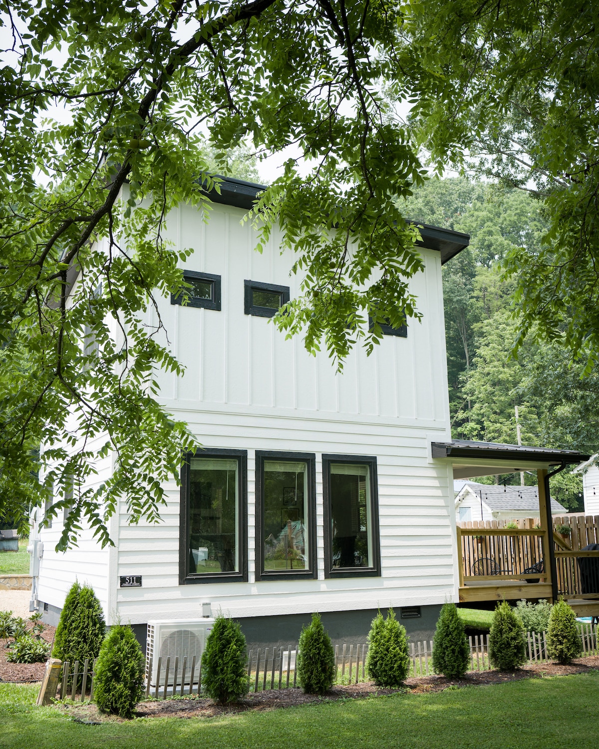 A modern two-story home is nestled among lush greenery, showcasing a white exterior with black accents. Large windows allow natural light to flood the interior, while a multi-level deck invites outdoor relaxation. Neatly trimmed hedges line the front, enhancing the inviting entrance.