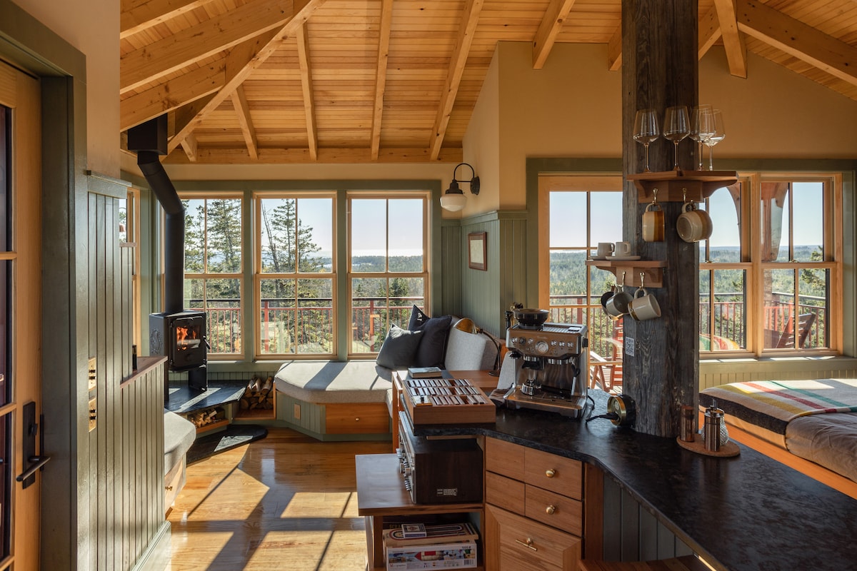 Natural light fills the airy interior space, highlighted by large windows offering expansive views. A cozy seating area features a wood stove, while a well-equipped kitchen includes a Breville espresso maker. Wooden beams and warm tones create a harmonious atmosphere.