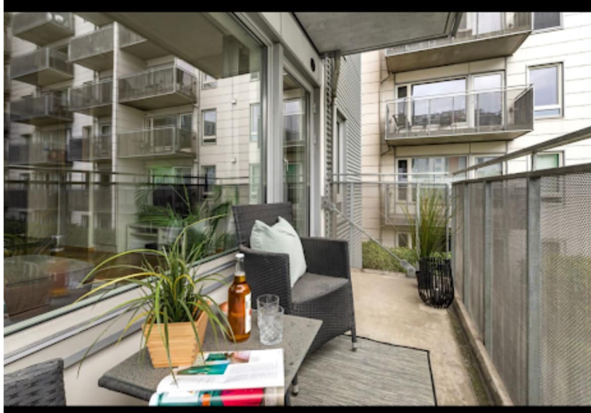 A private balcony features two woven chairs positioned beside a small table. A potted plant adds a touch of greenery. Large glass doors provide views into the building's courtyard, which includes multiple apartment balconies and a quiet, spacious atmosphere.