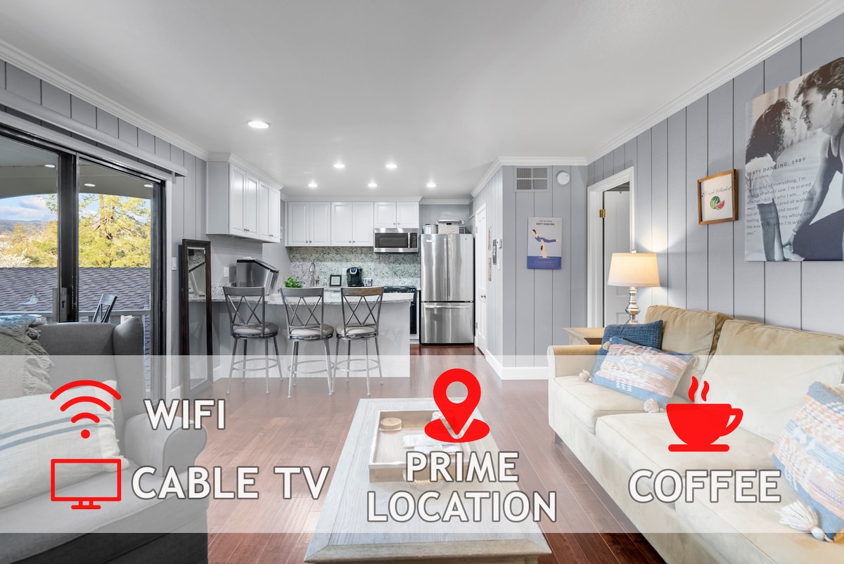 An open living area is presented, featuring a comfortable sofa and a dining table with high-backed chairs. The kitchen is equipped with modern appliances, and large glass doors lead to an outdoor space, allowing natural light to fill the room.