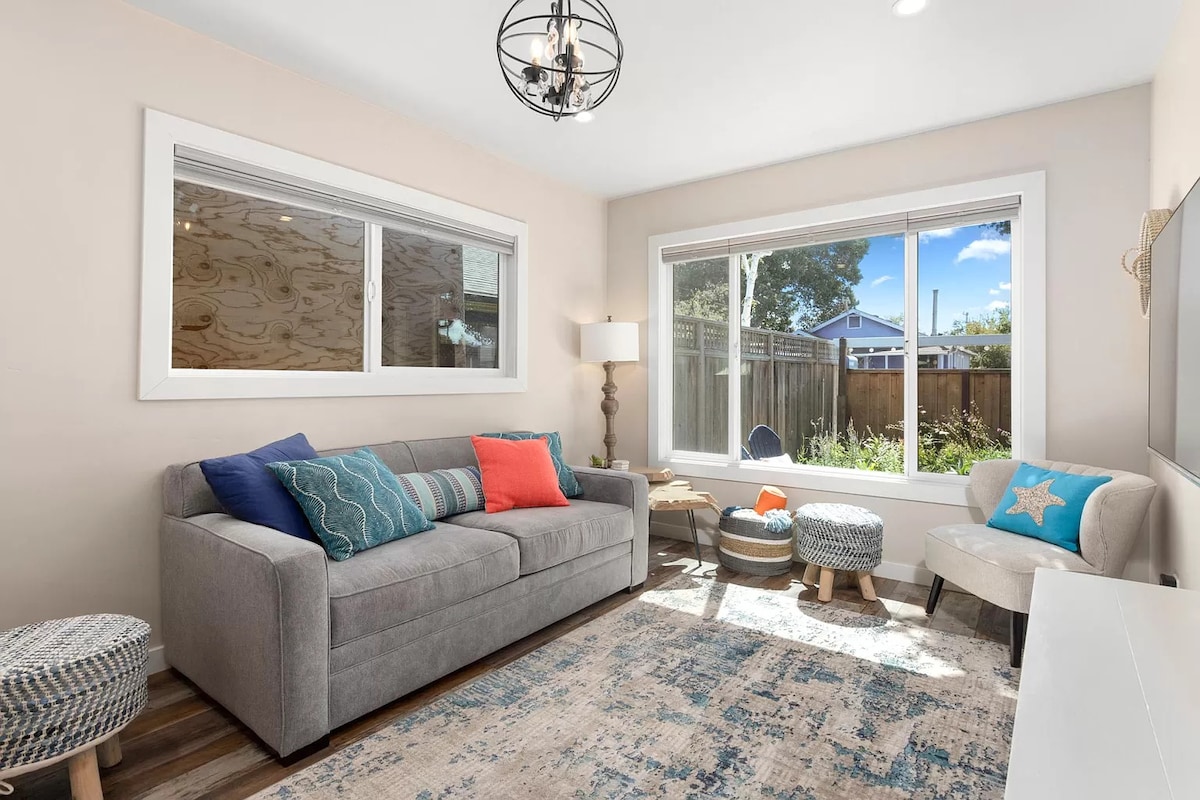 A bright living area features a comfortable gray sofa adorned with colorful cushions. Natural light pours in through large windows, illuminating a patterned rug and enhancing the space's inviting feel. A stylish lamp stands beside the sofa, complementing the overall decor.