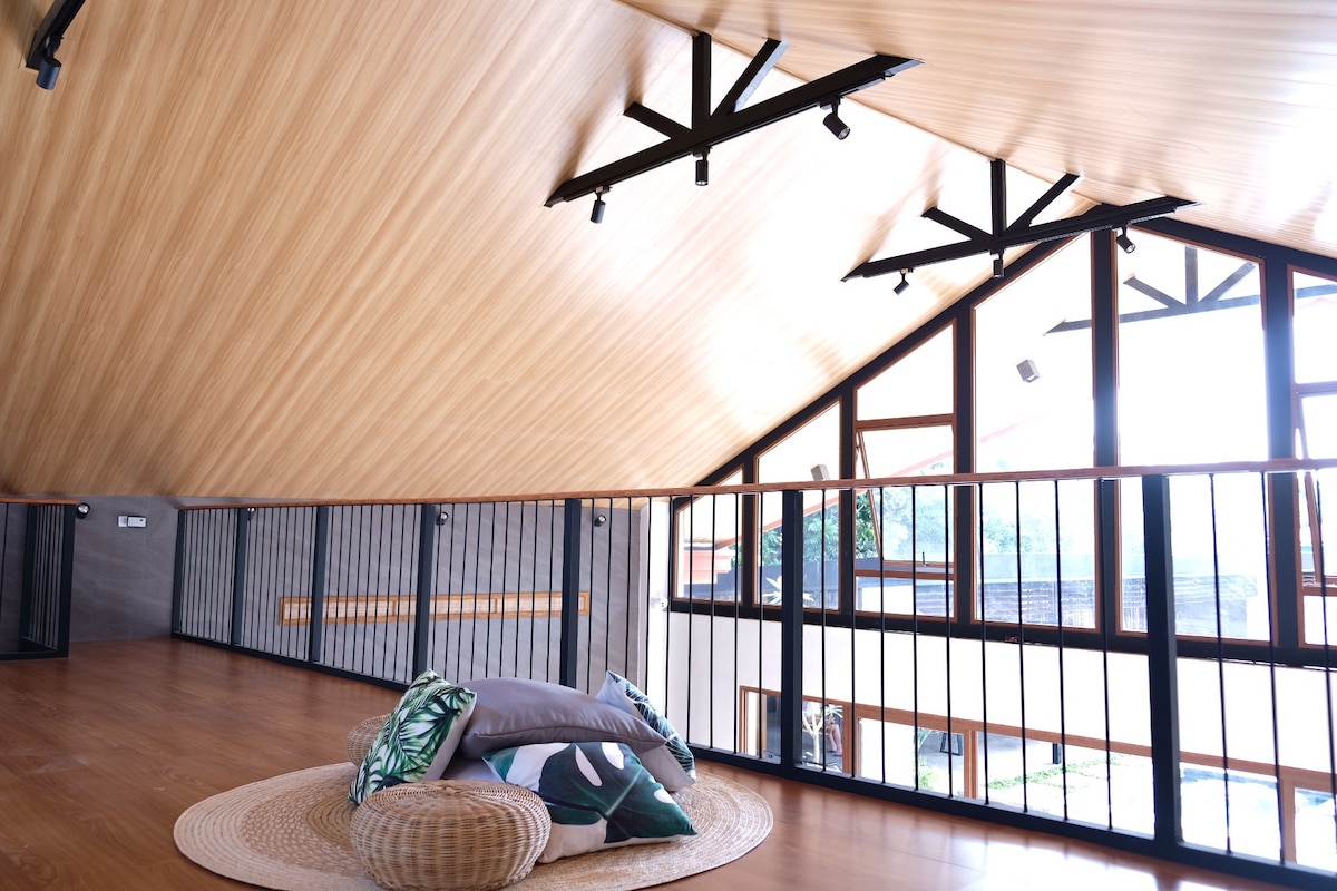 An open loft space features a wooden ceiling with exposed beams and modern lighting fixtures. A circular area rug is placed on the polished wooden floor, surrounded by cushions in varying shades of green and grey. Large windows allow natural light to fill the room.
