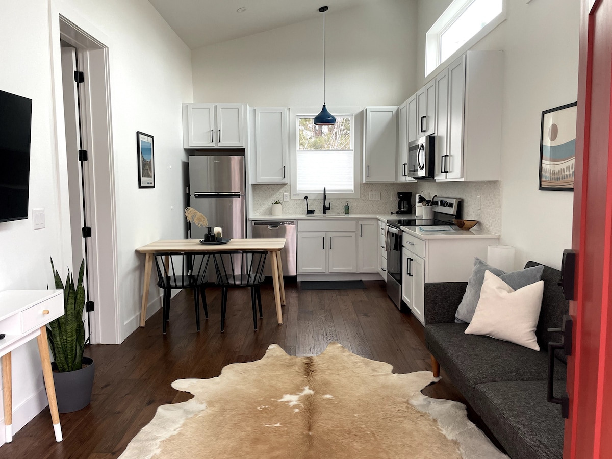 An open floor plan kitchen and living area showcases modern appliances and furnishings. A dining table sits adjacent to the kitchen, while a cozy seating area includes a gray couch with decorative pillows. A light-colored area rug complements the wooden flooring.
