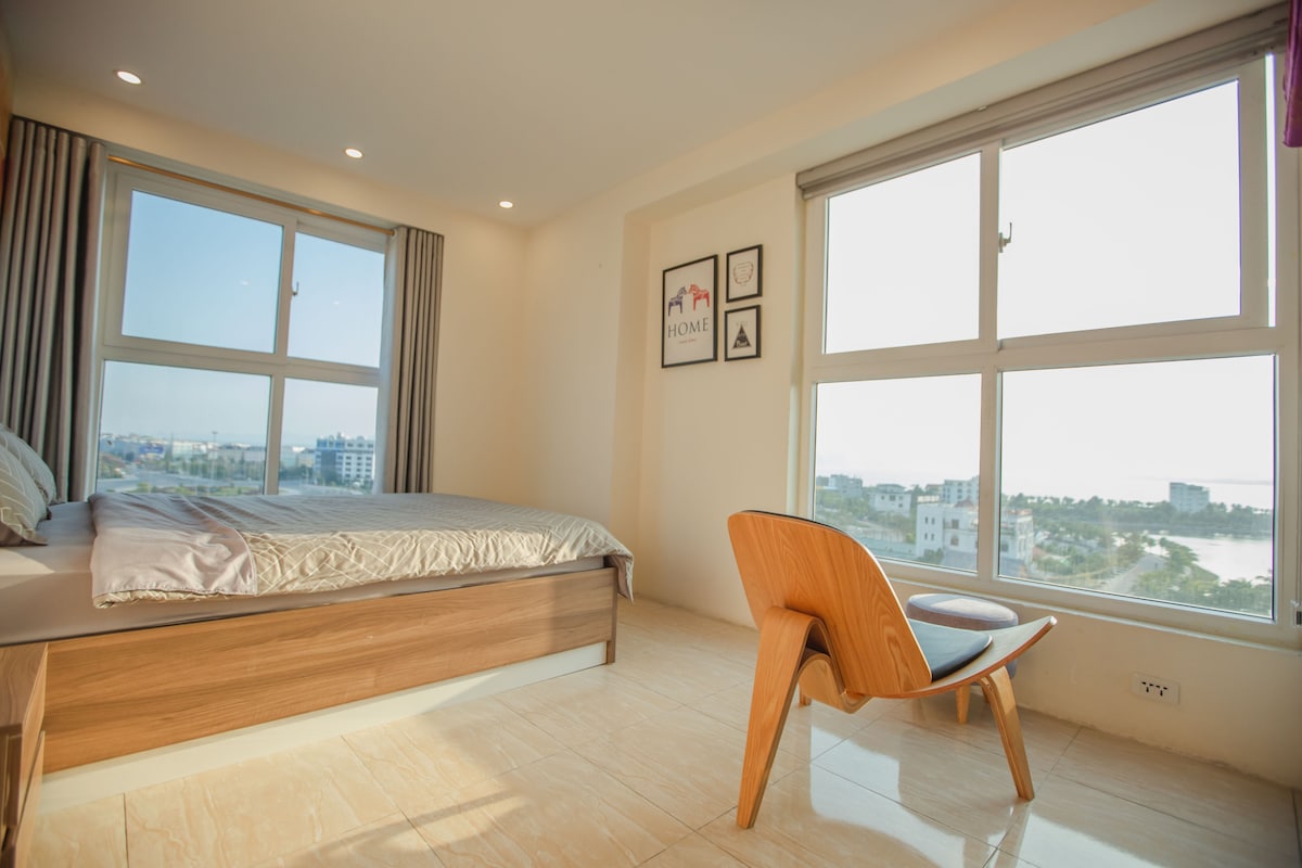 A spacious bedroom features large windows offering stunning views of the surrounding area. A comfortable bed is accompanied by a sleek wooden chair, creating a simple yet inviting atmosphere. Natural light fills the room, enhancing the clean and modern design.
