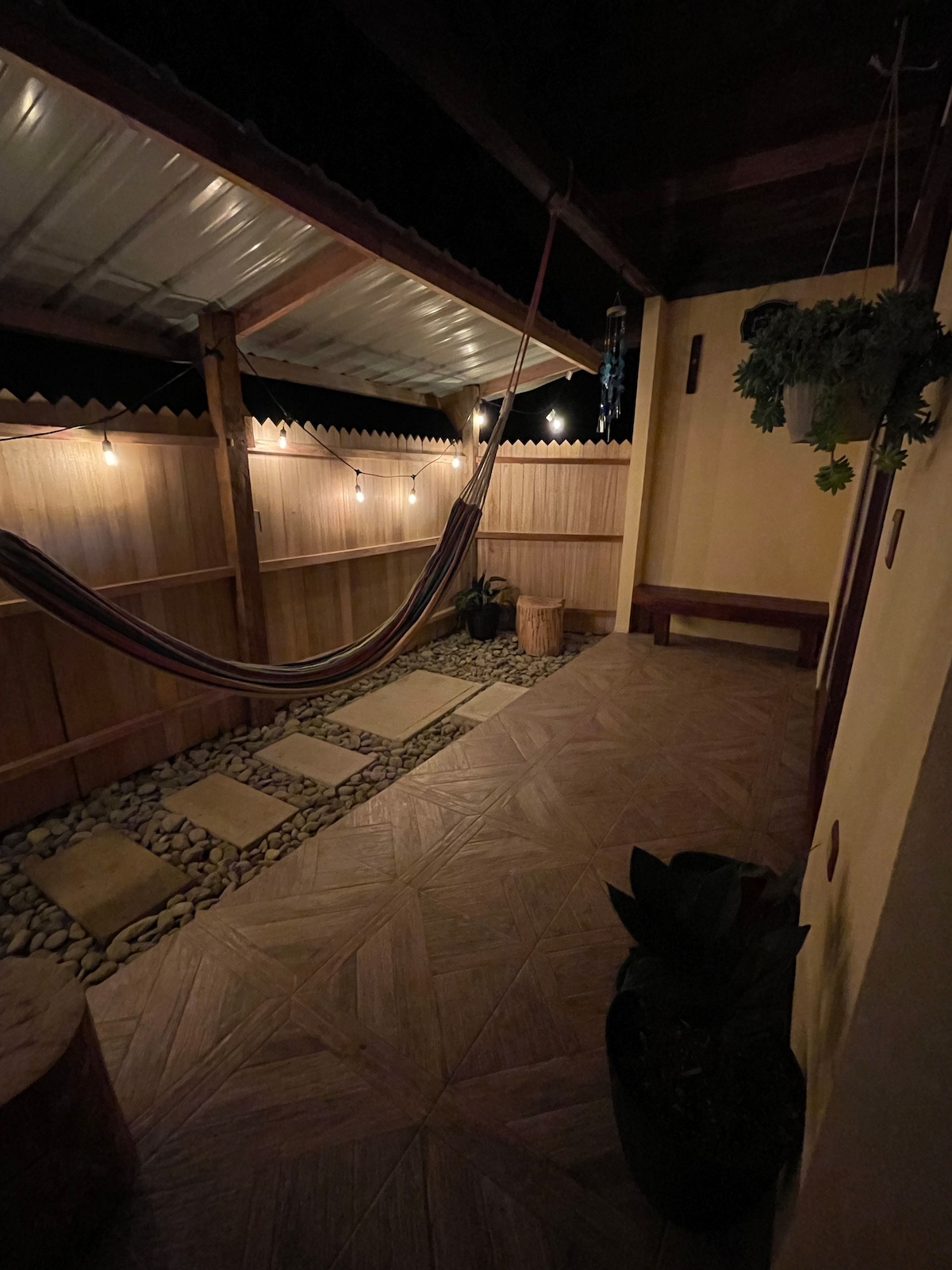 An outdoor space is illuminated by soft string lights, featuring a hammock suspended between wooden beams. A pathway of stones leads to a well-maintained garden with potted plants. The area is bordered by illuminated wooden fencing, creating a cozy atmosphere.