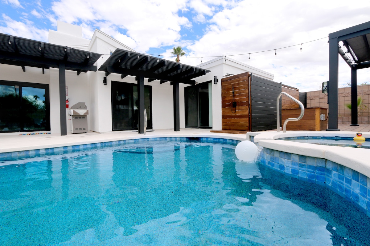 A clear view of the inviting pool surrounded by modern architecture is presented. The pool features a gentle entry step and a buoy floating on the surface. Nearby, a shaded outdoor seating area is visible, with a grill and wooden partitions enhancing the space.