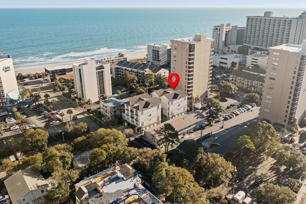An aerial view showcases the duplex structure surrounded by trees, with the ocean in the background. The listing is highlighted by a red marker, indicating its central location near the beach and nearby buildings, demonstrating the proximity to surrounding amenities.