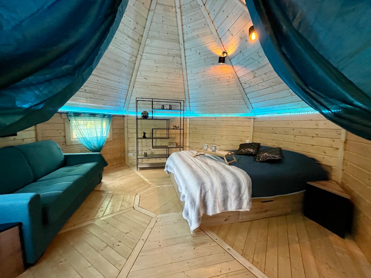 A circular room features wooden walls and a hexagonal floor, with a spacious bed at the center. A comfortable couch and a small shelf are present. Soft lighting is provided by hanging bulbs, while sheer curtains allow natural light to filter in.