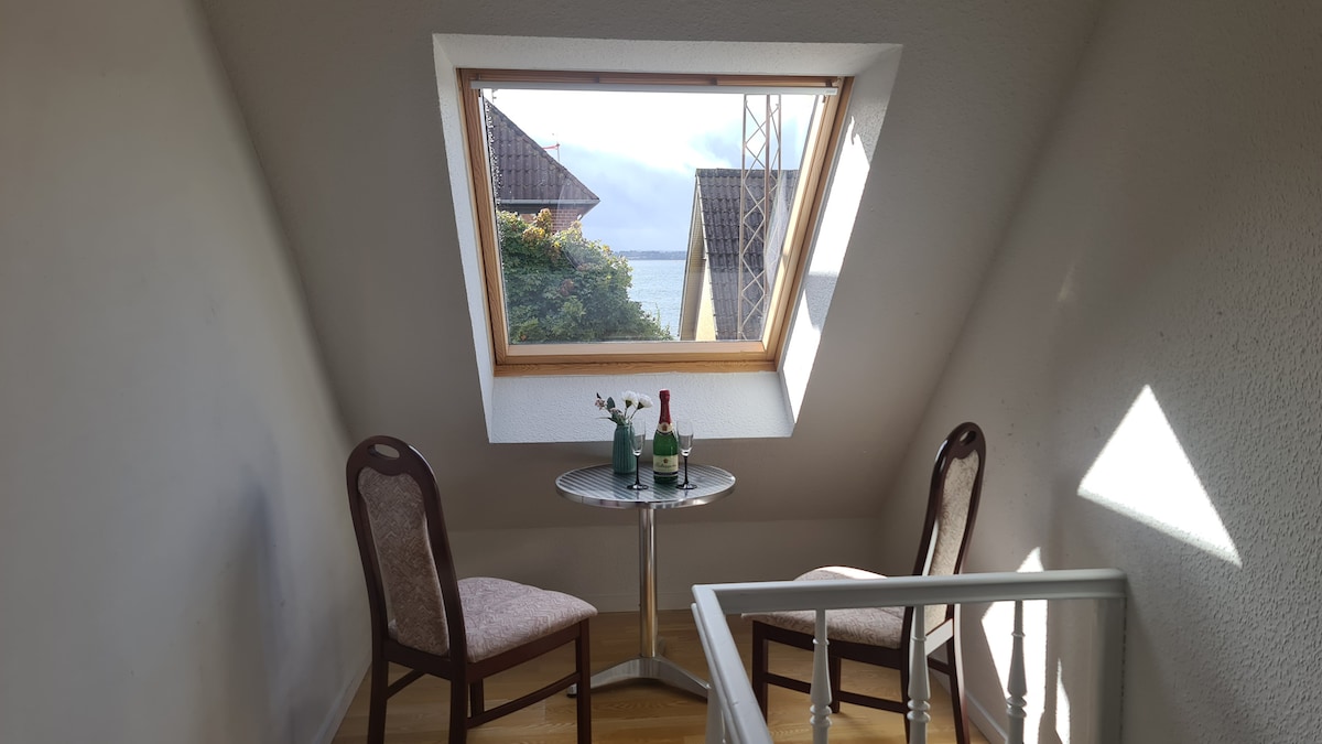 A cozy nook features a small round table set for two, accompanied by two wooden chairs. A bottle of sparkling wine and two glasses are positioned centrally, while sunlight filters in through a triangular window, offering a view of the surroundings.