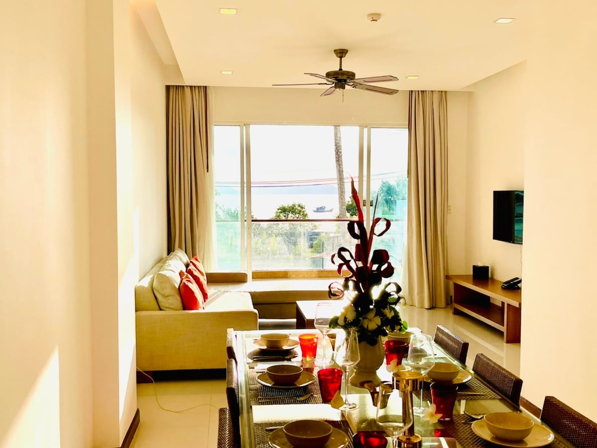 A light-filled living area presents a dining table set for four, featuring red glassware and plate ware. A comfortable sofa with accent pillows faces large windows, which offer views of the sea. A ceiling fan and entertainment unit are also visible.